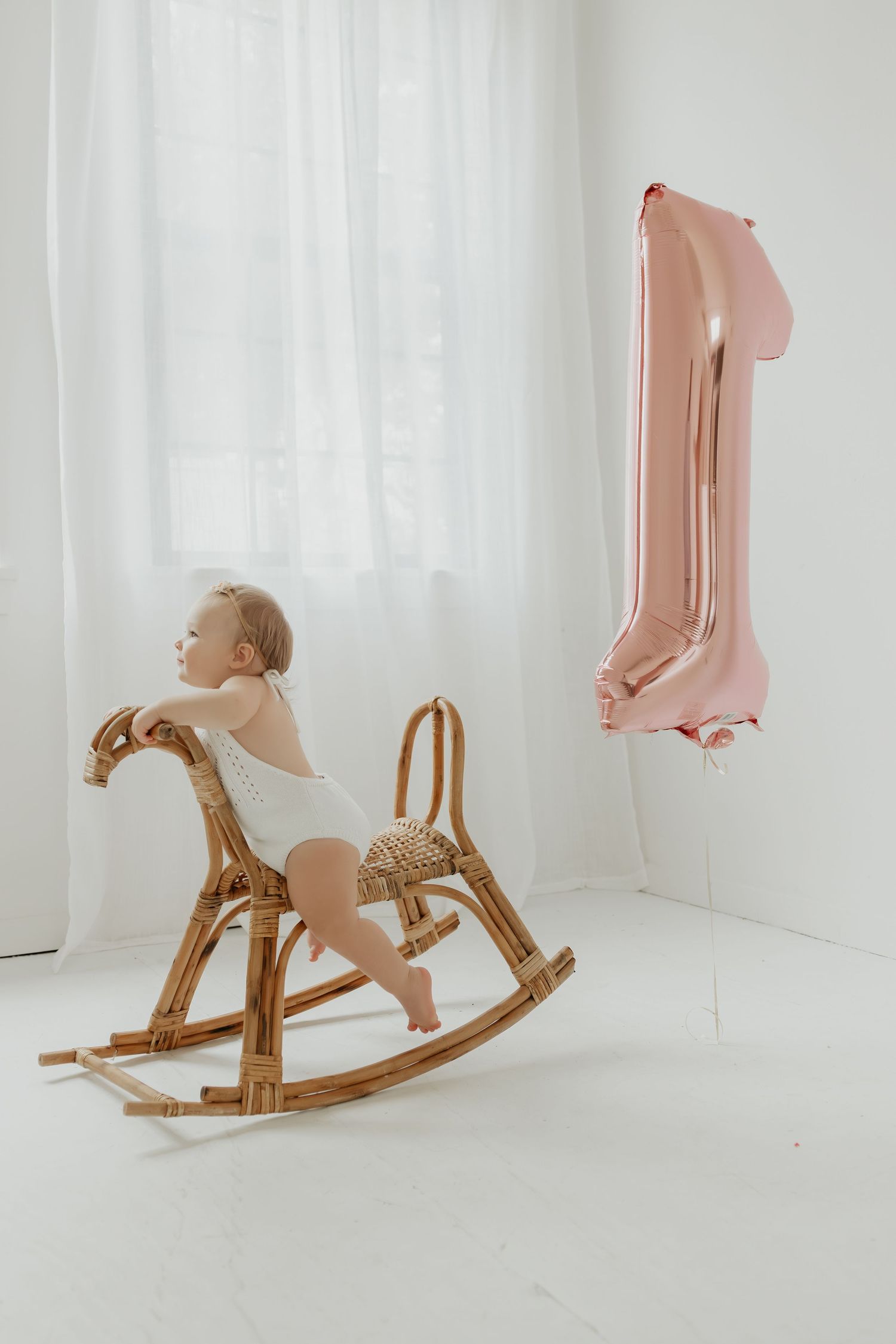 Baby riding wooden rocking horse with pink number one balloon in white studio setting.