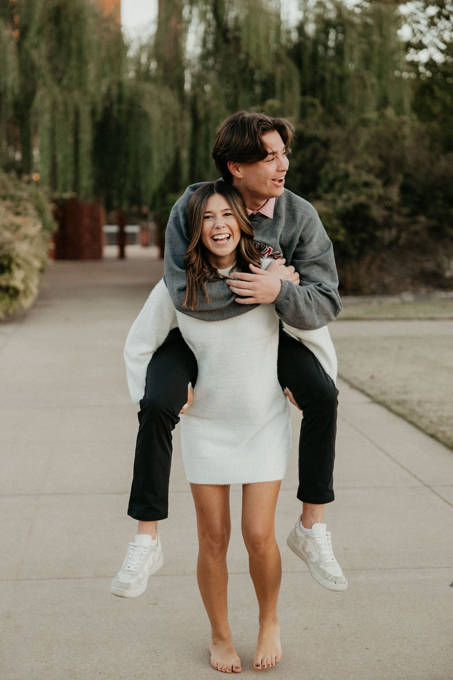 Playful couple in casual wear having fun on a sidewalk with trees in background.