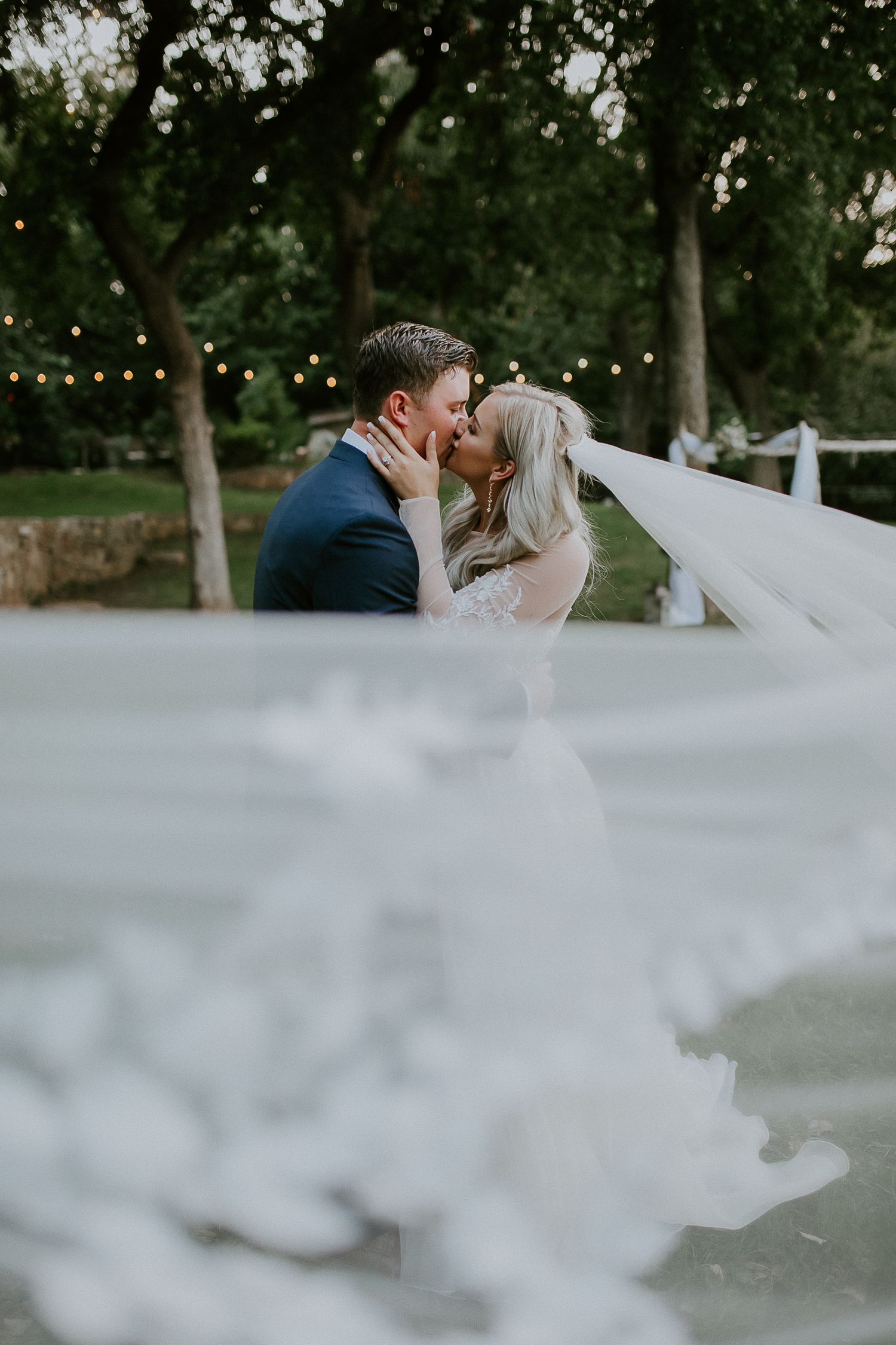 Romantic outdoor wedding scene with flowing veil and string lights in garden setting.