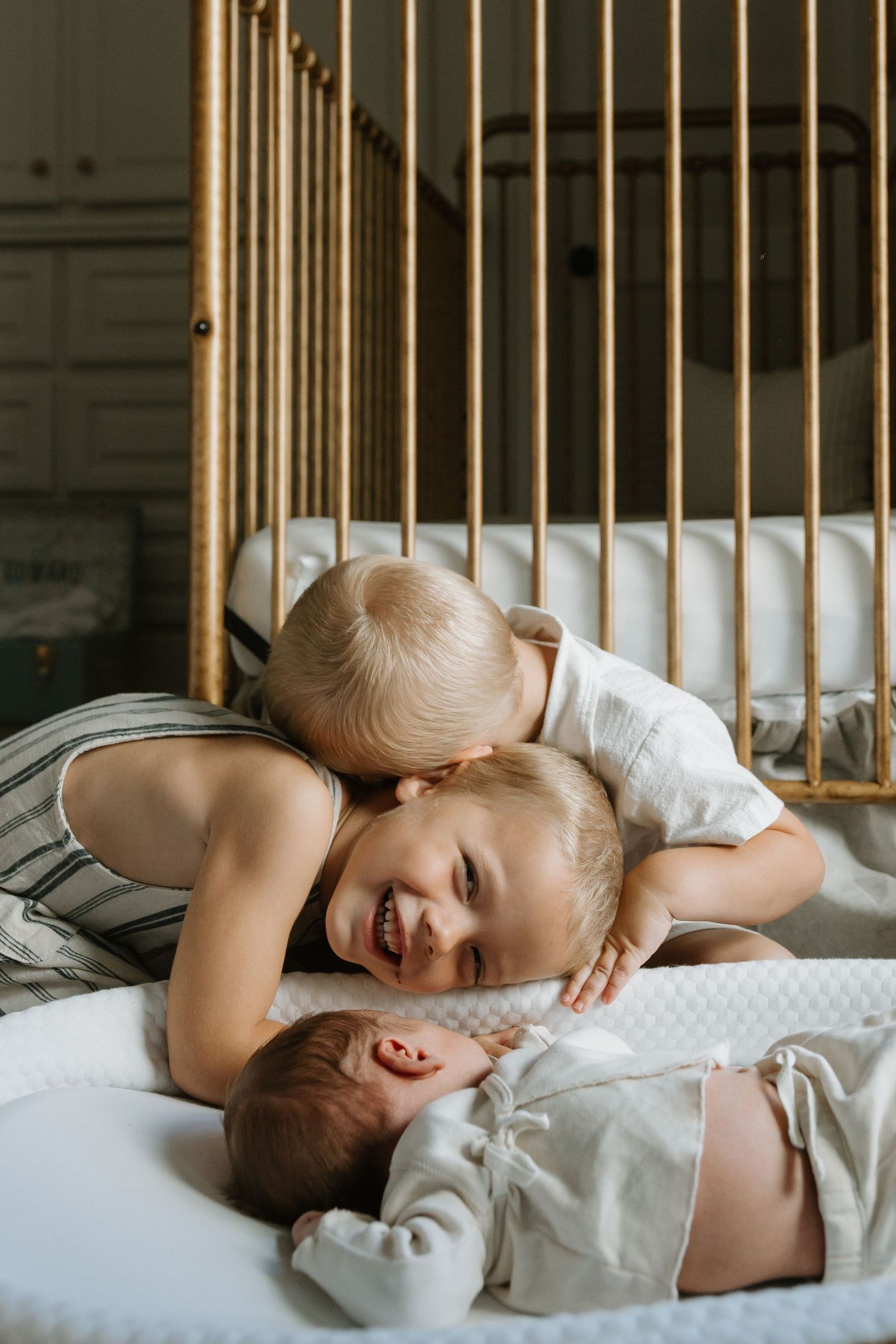 Two young children resting together on white bedding with golden crib rails in background.