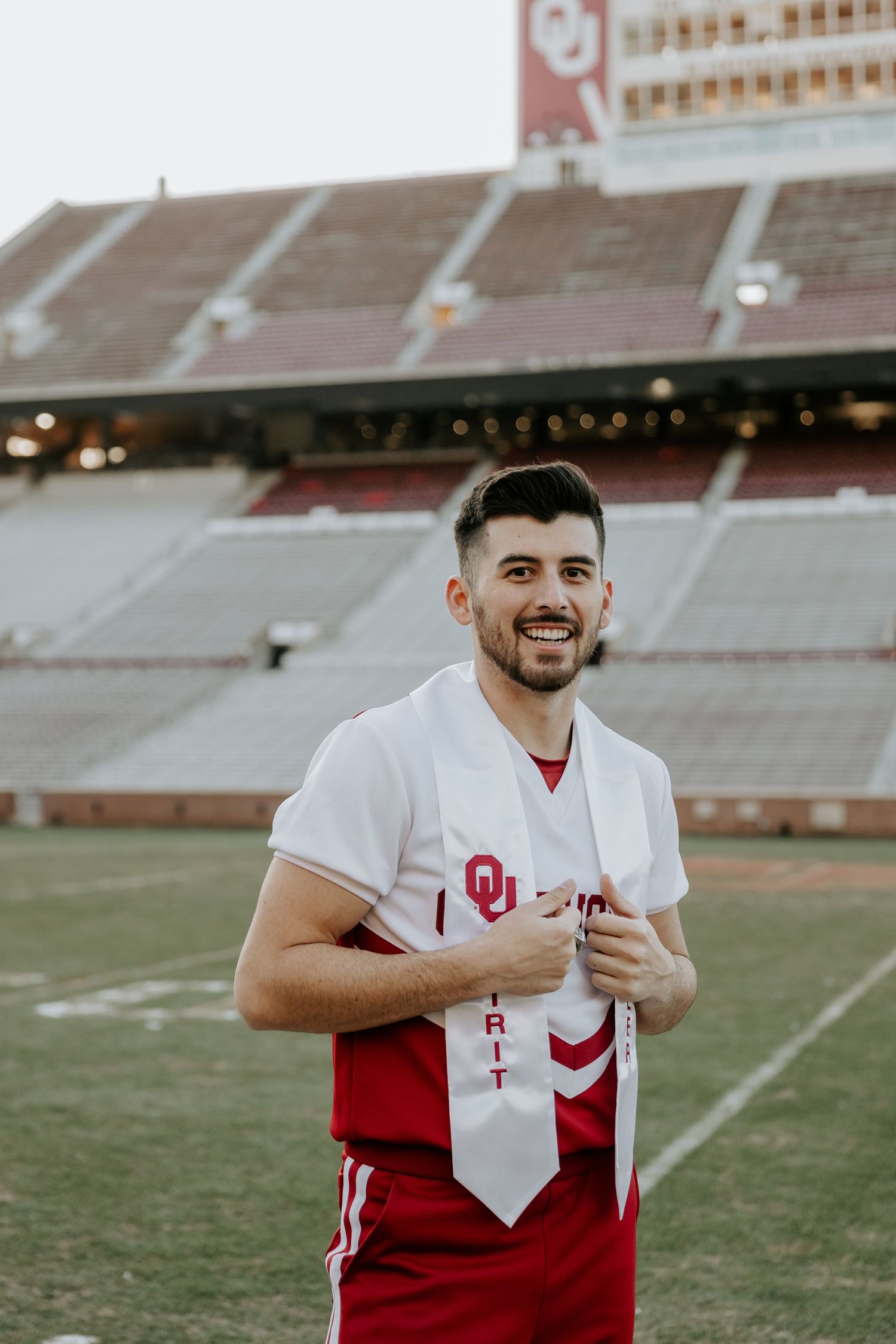 Graduate wearing red outfit and white stole stands proudly on OU football field.