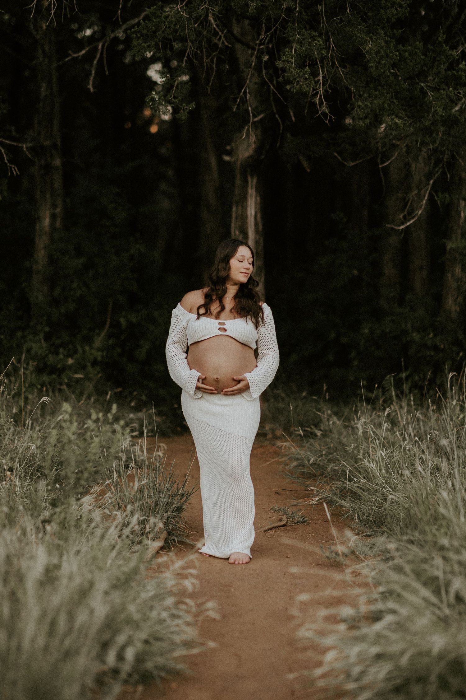 Moody maternity photos in white dress showing silhouette against dark forest background.