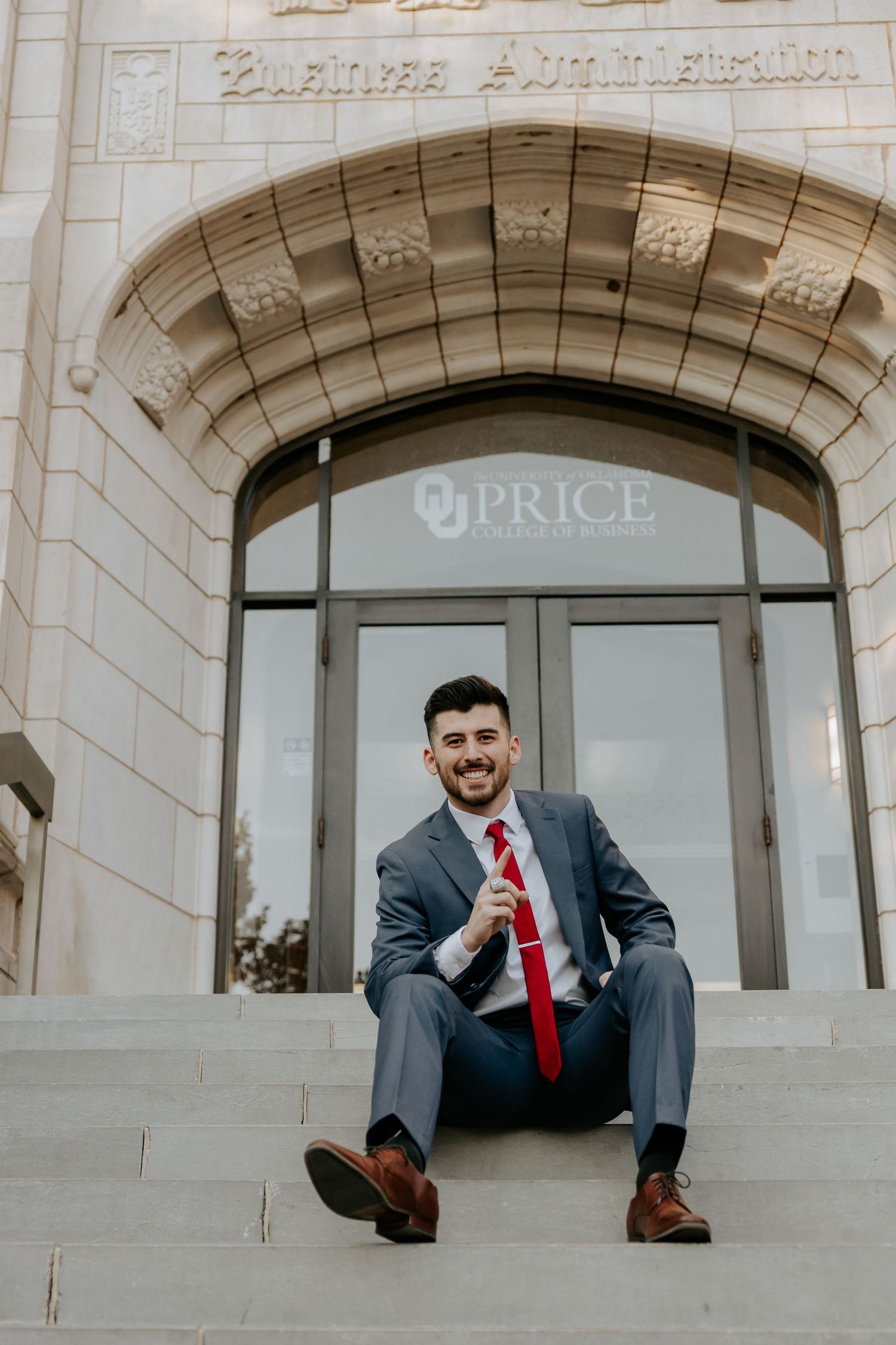 Graduate in business attire sits on stone steps of university building during photo session.