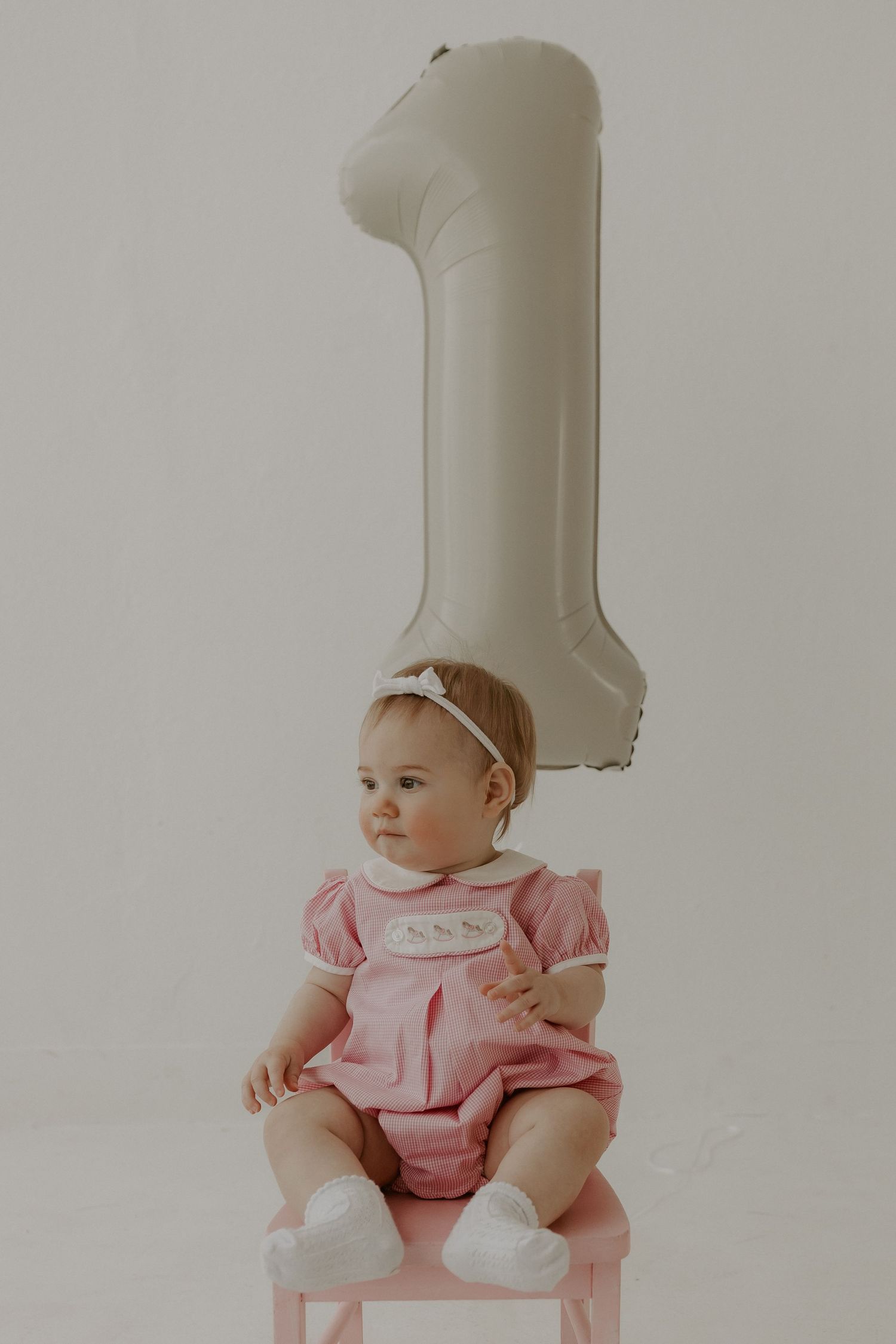 Baby in pink outfit sitting with large number 1 balloon.
