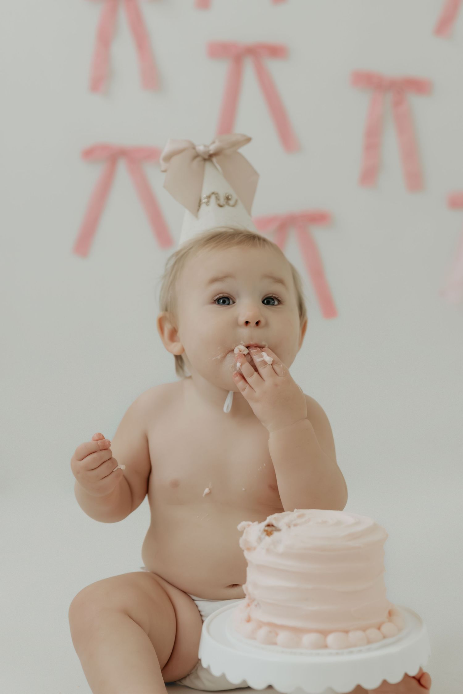 Pink themed cake smash photography session with white frosted cake and bow decorations.