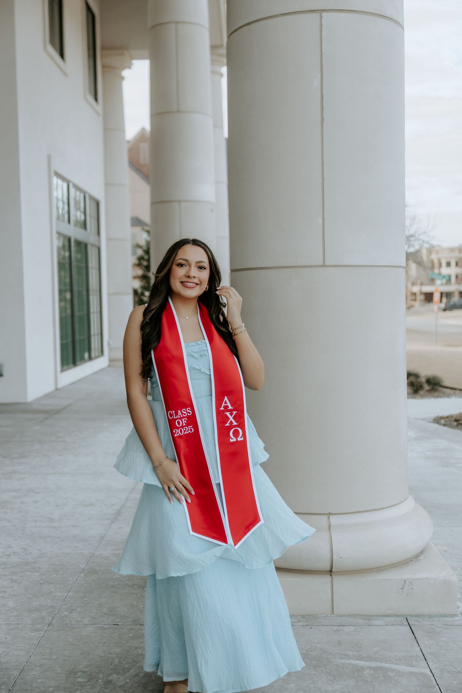 Graduate in a light blue dress and red stole poses between white columns during a graduation photoshoot.