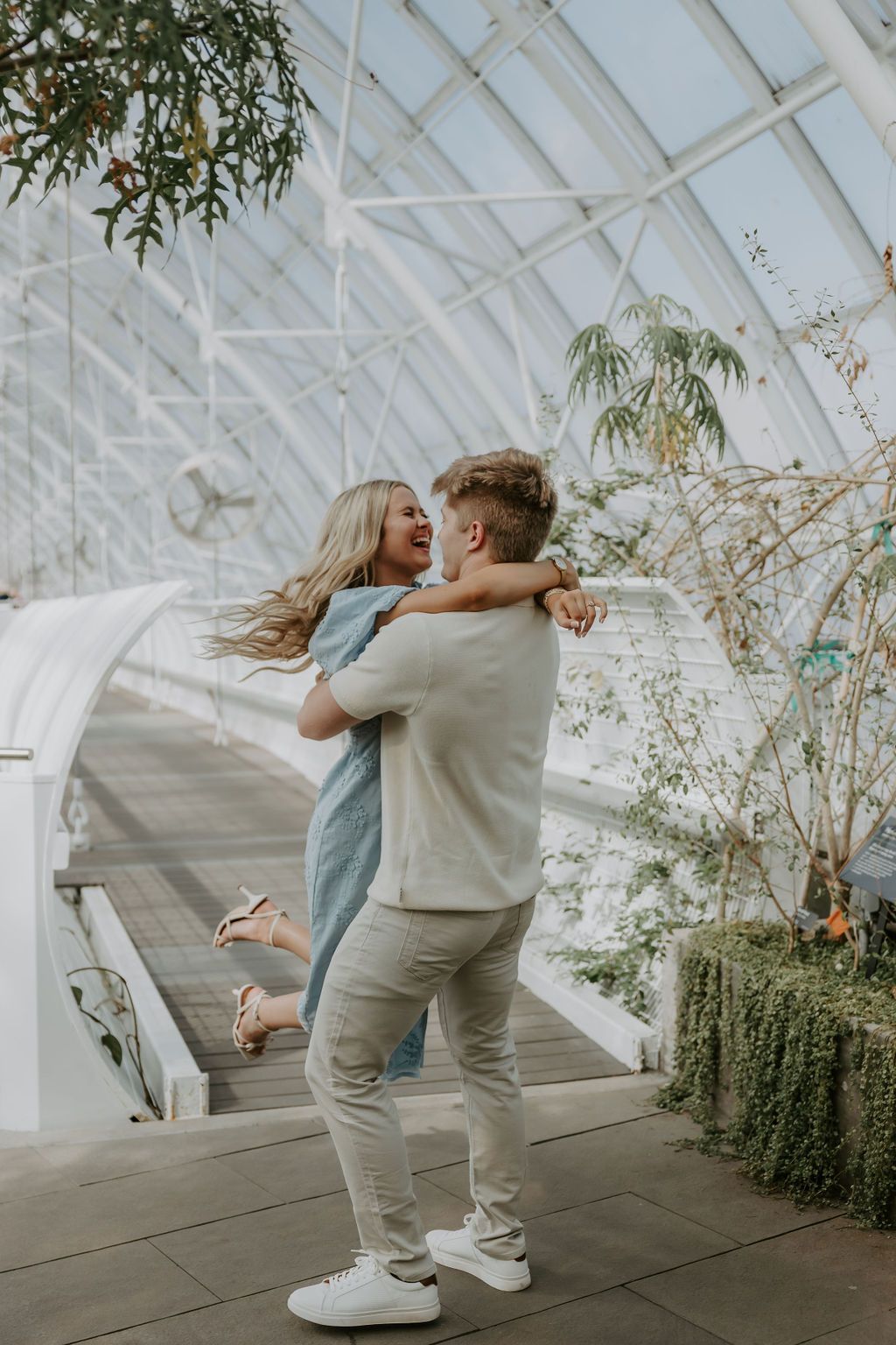 Dancing couple in casual outfits enjoying playful moments in sunlit greenhouse.