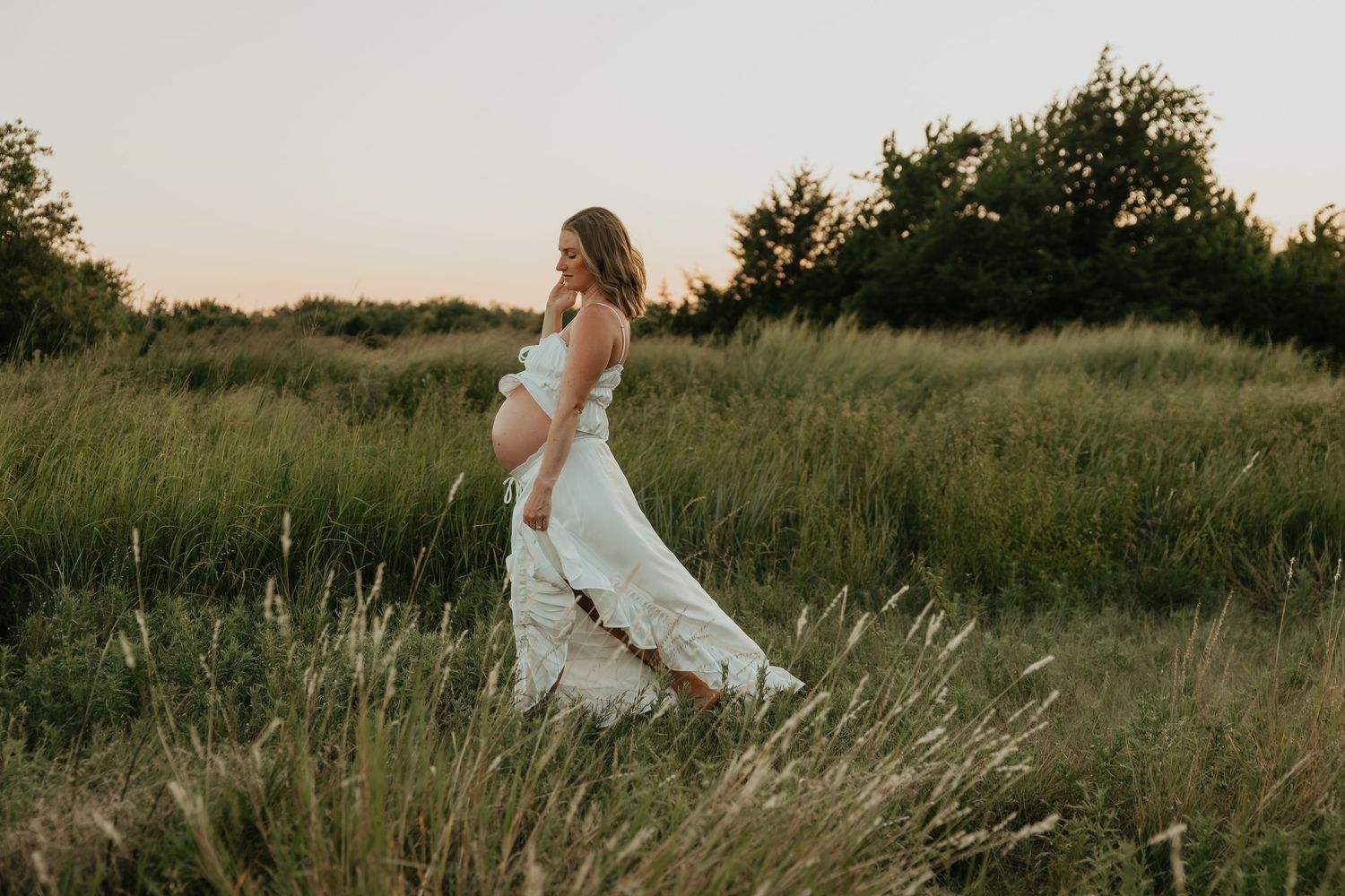 A white dress flows in the breeze as someone poses in a serene meadow at sunset.