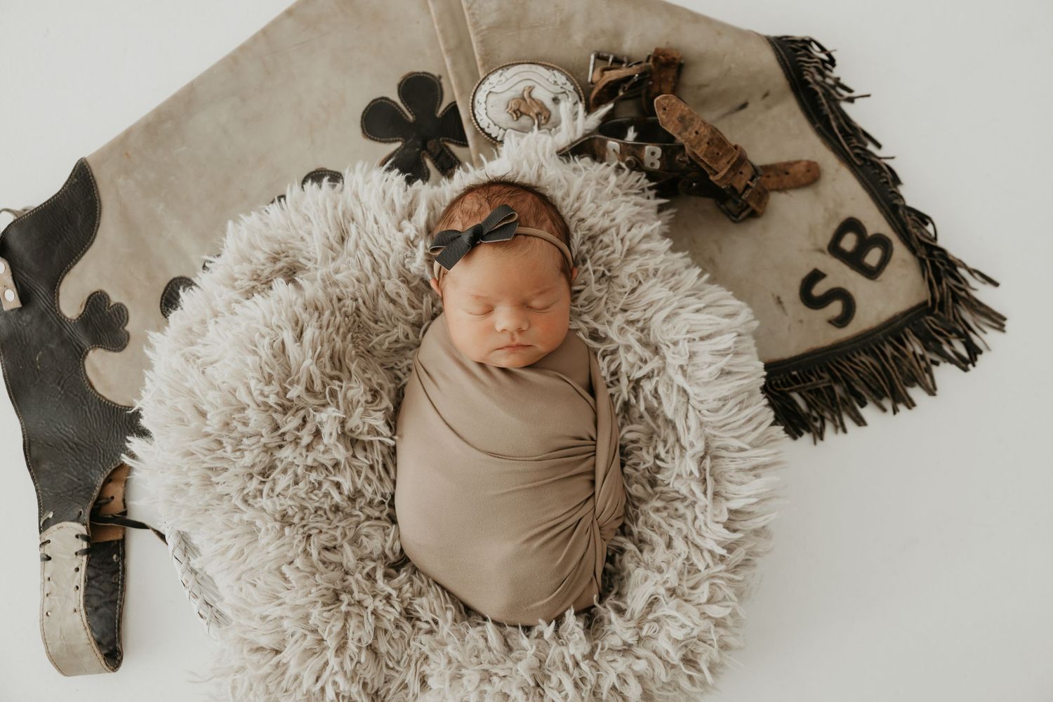 Newborn baby wrapped in beige swaddle lying on textured rug with decorative accessories.