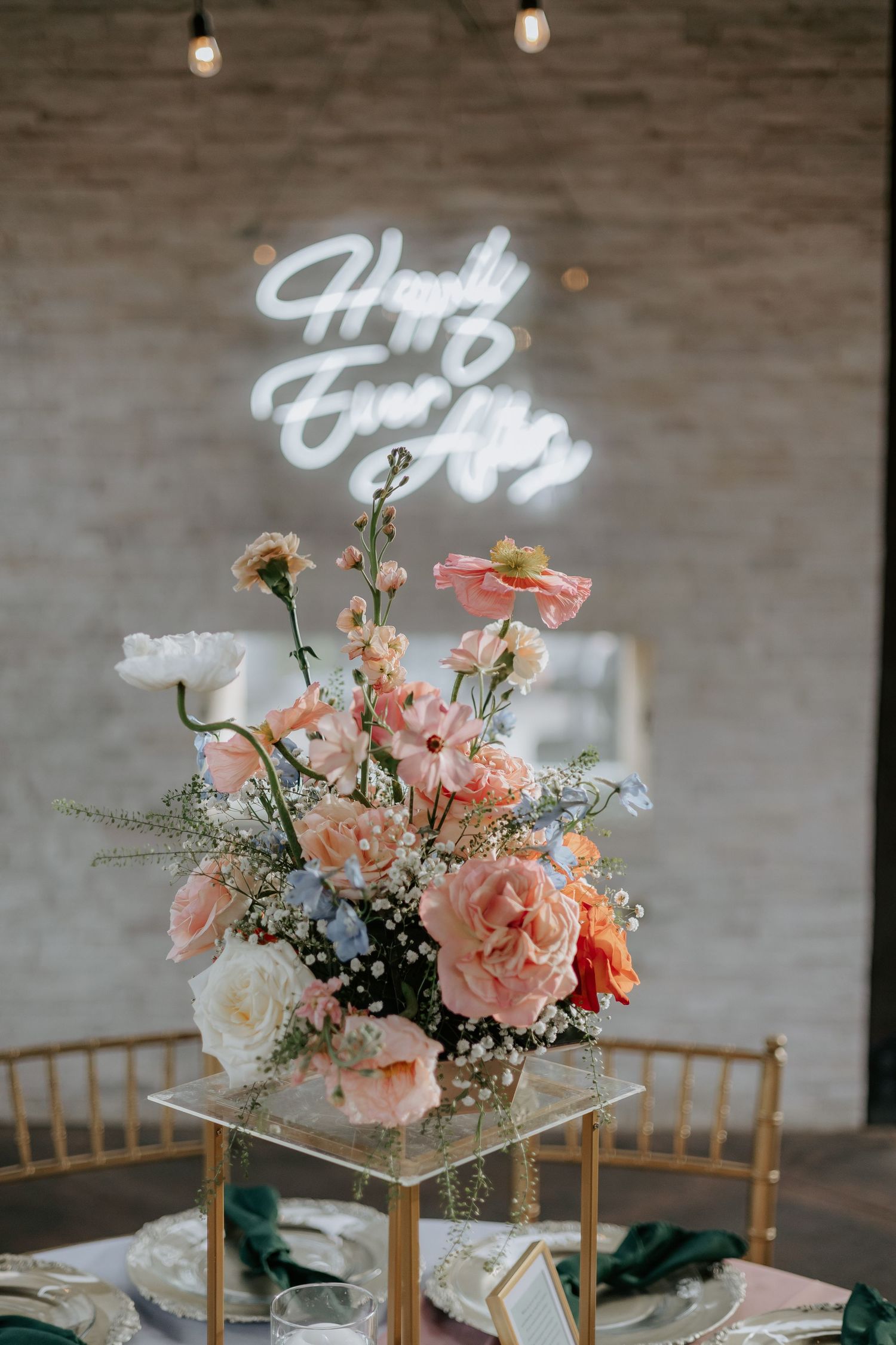 Elegant floral centerpiece with pink blooms and greenery against brick wall with illuminated signage.