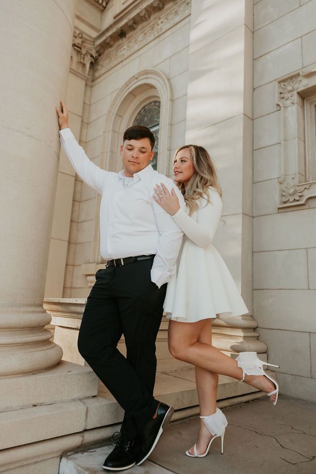 Stylish couple in white outfits posing playfully against classical architecture.