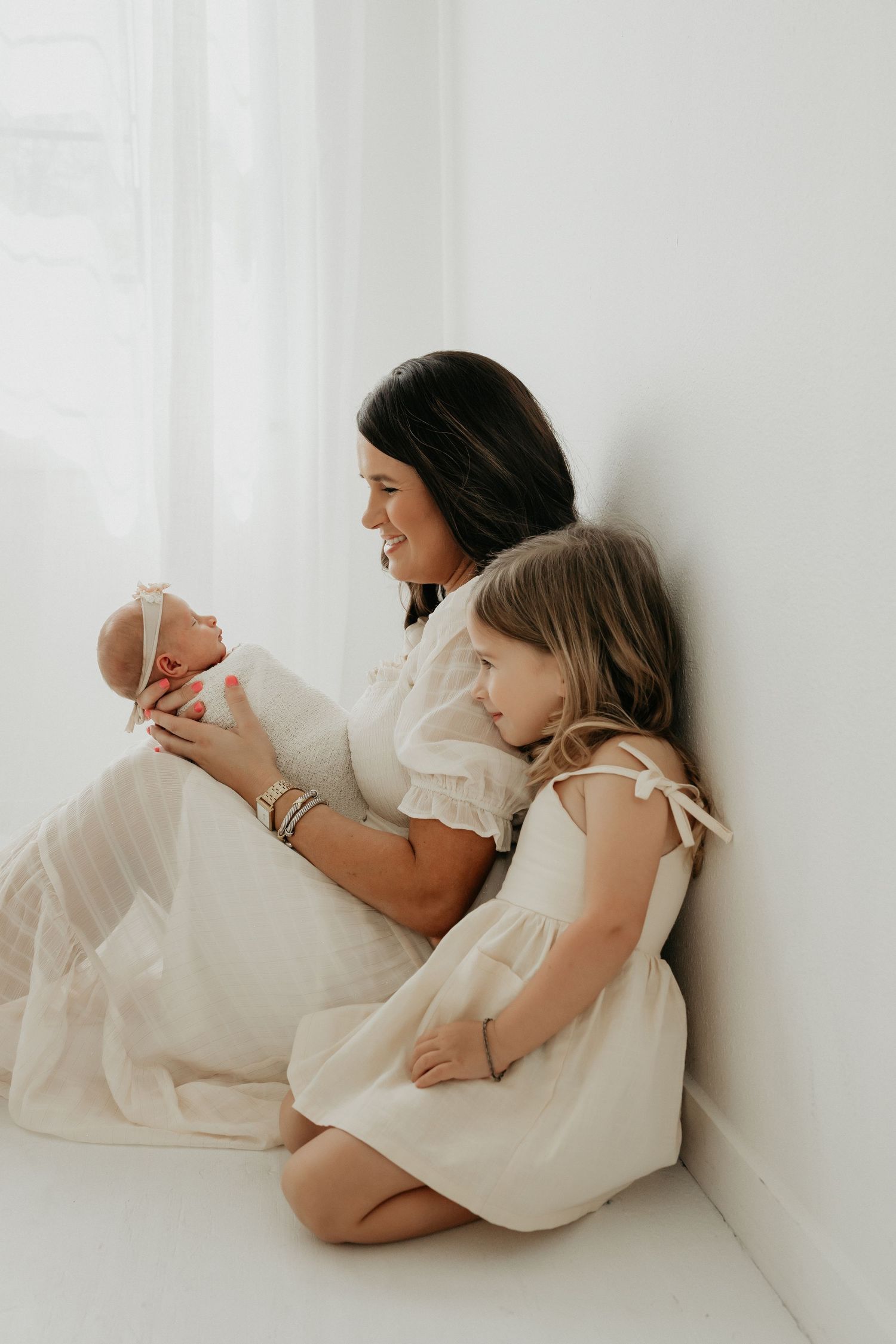 Mother and child in white dresses resting together against white wall.