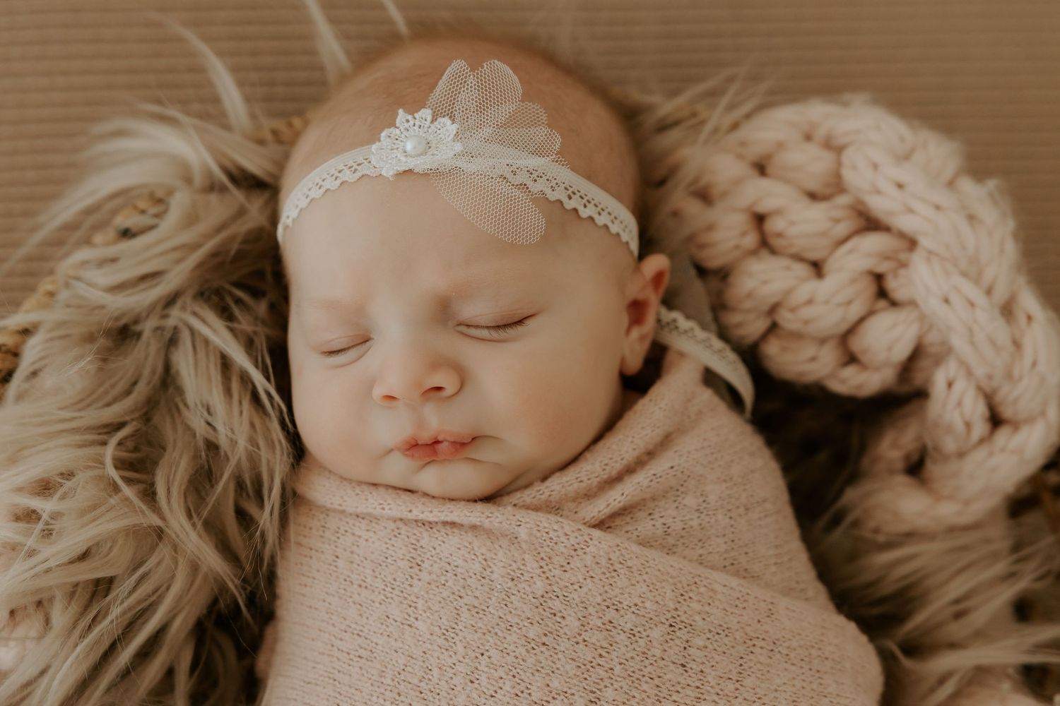 Newborn resting on textured beige blanket with delicate headpiece.