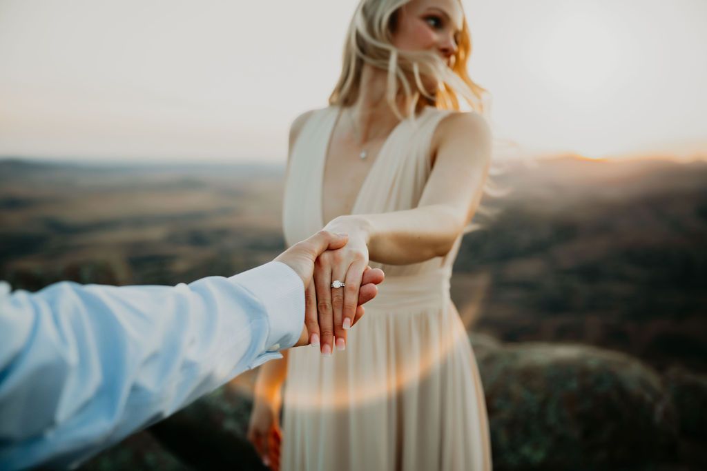 A couple holds hands in a white dress against a scenic mountain backdrop at sunset.