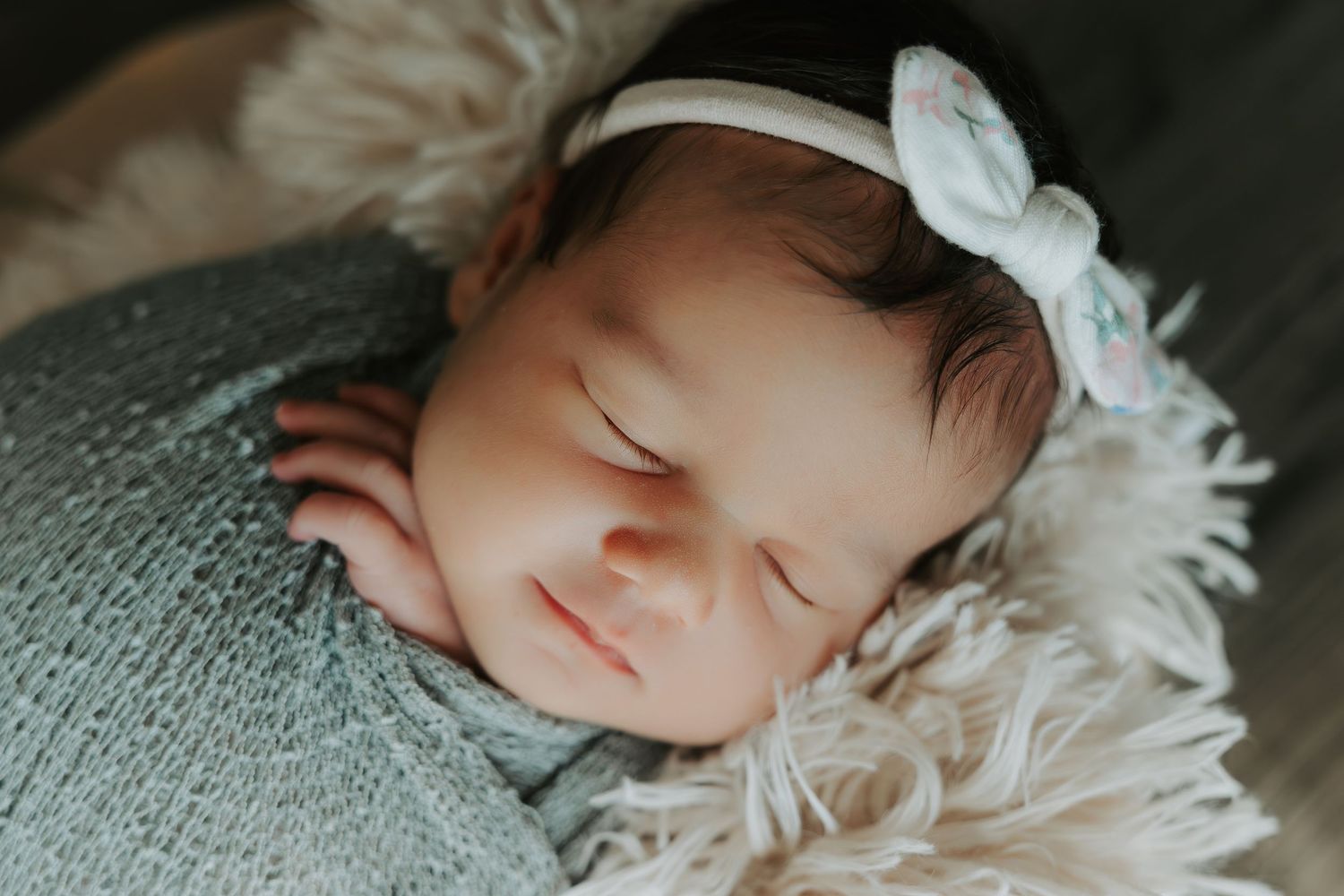 Sleeping newborn with white bow accessory resting on gray textured surface.