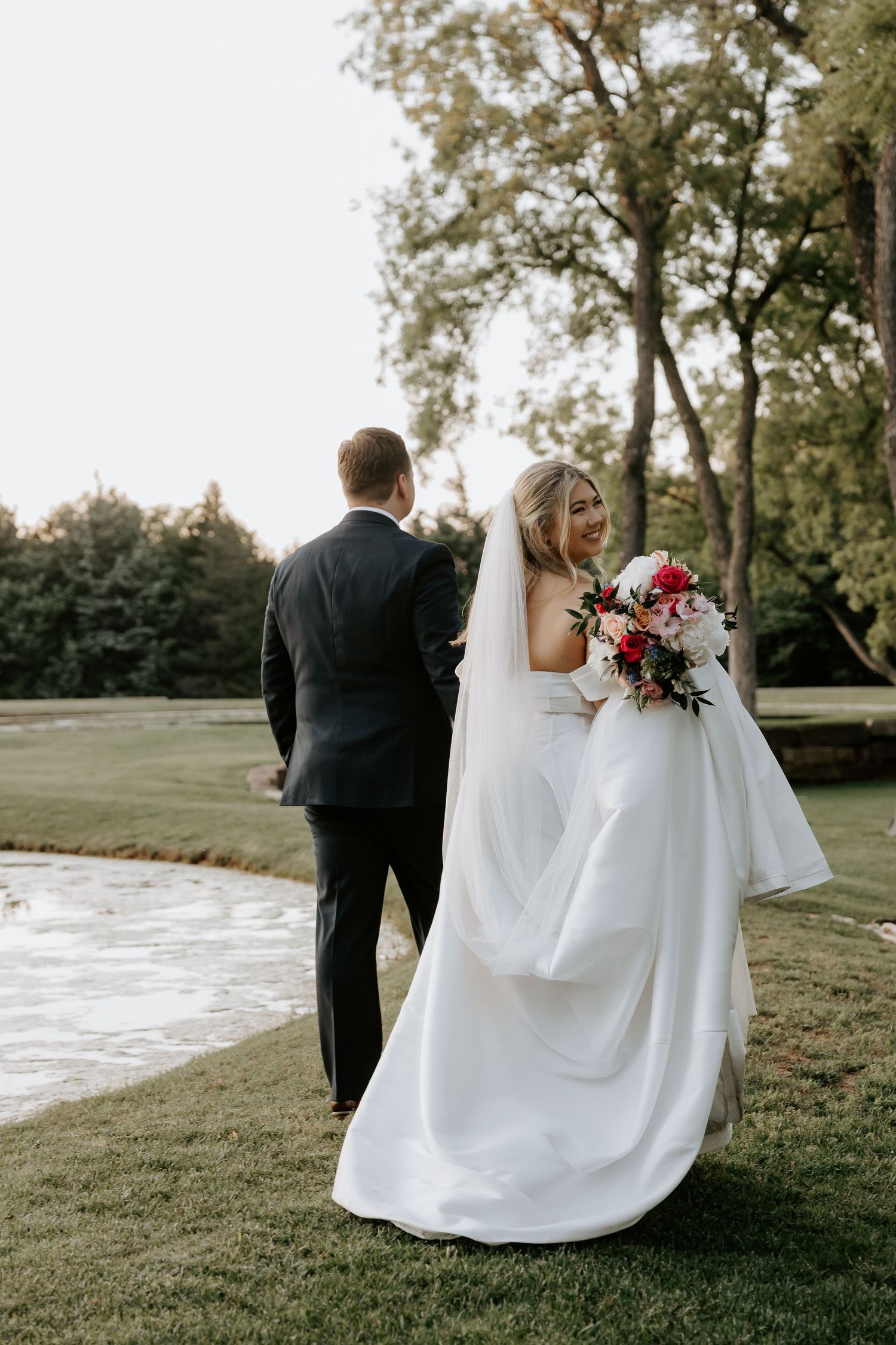 Couple walks along grassy path near water in wedding attire.