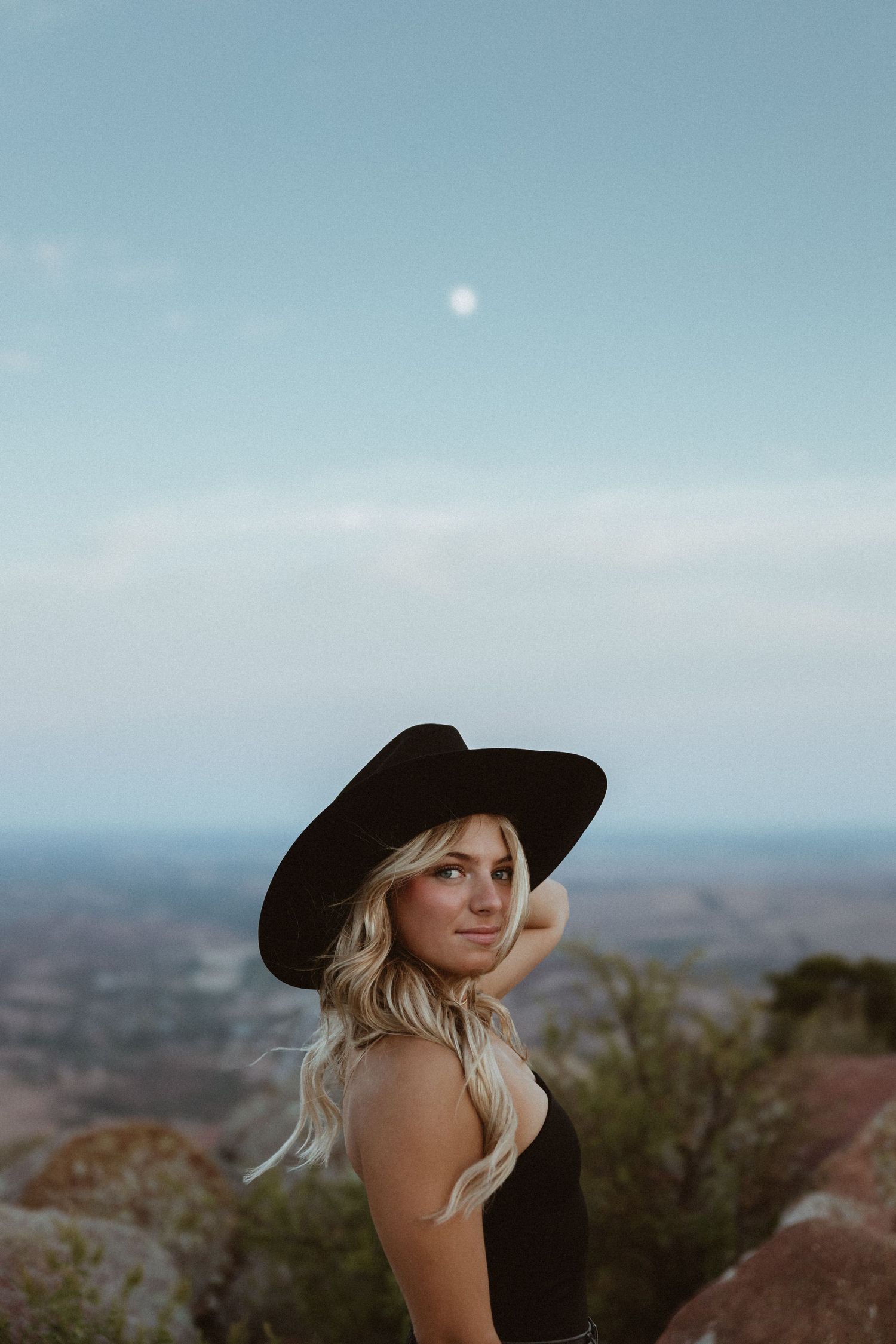 Person in black hat poses against desert landscape during sunset.