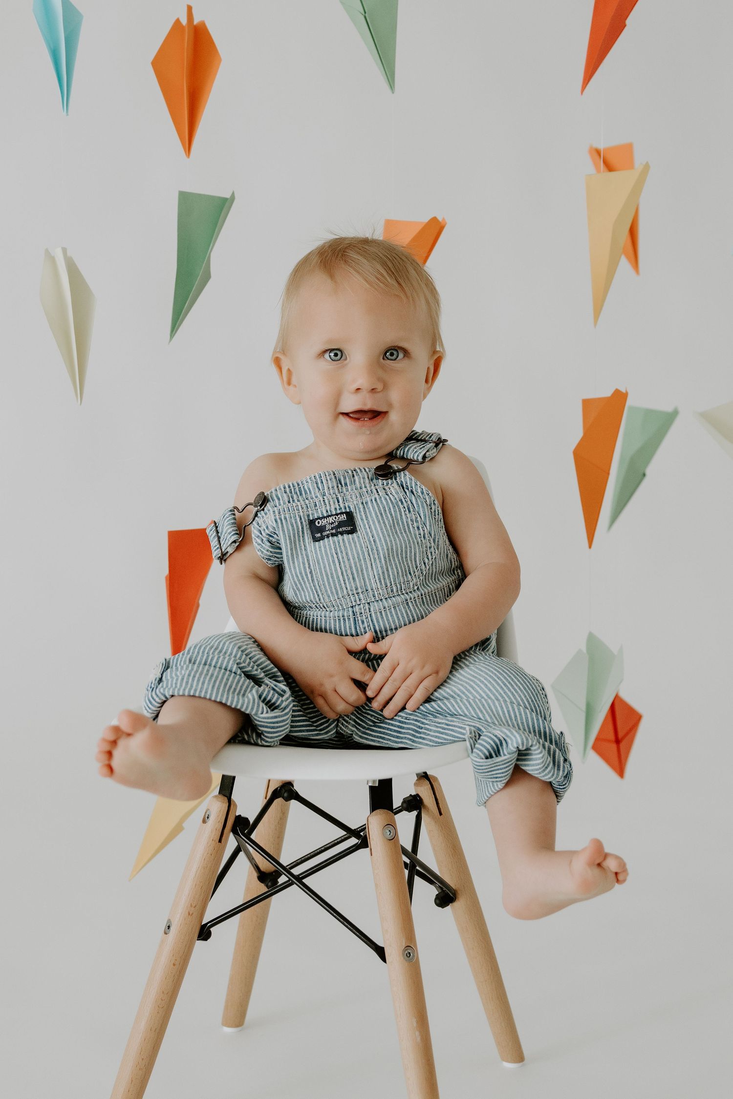 Baby in striped overalls sitting on modern chair with colorful paper decorations.