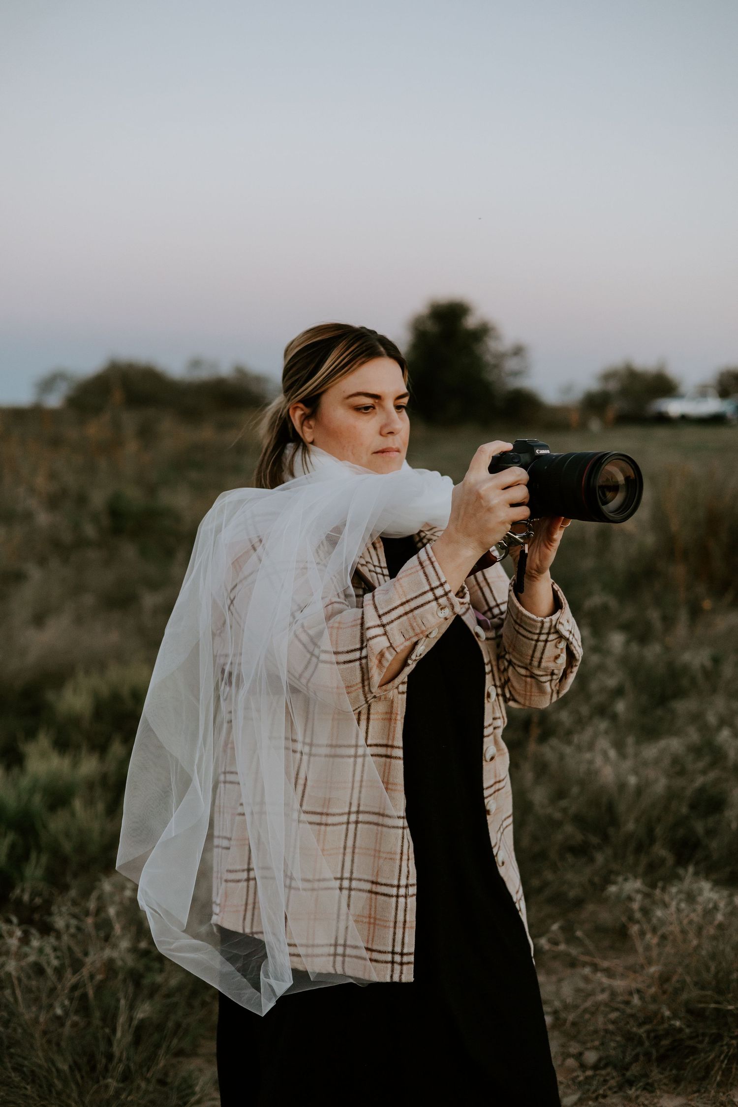 Photographer in plaid shirt and flowing veil holds professional camera in outdoor field during golden hour.