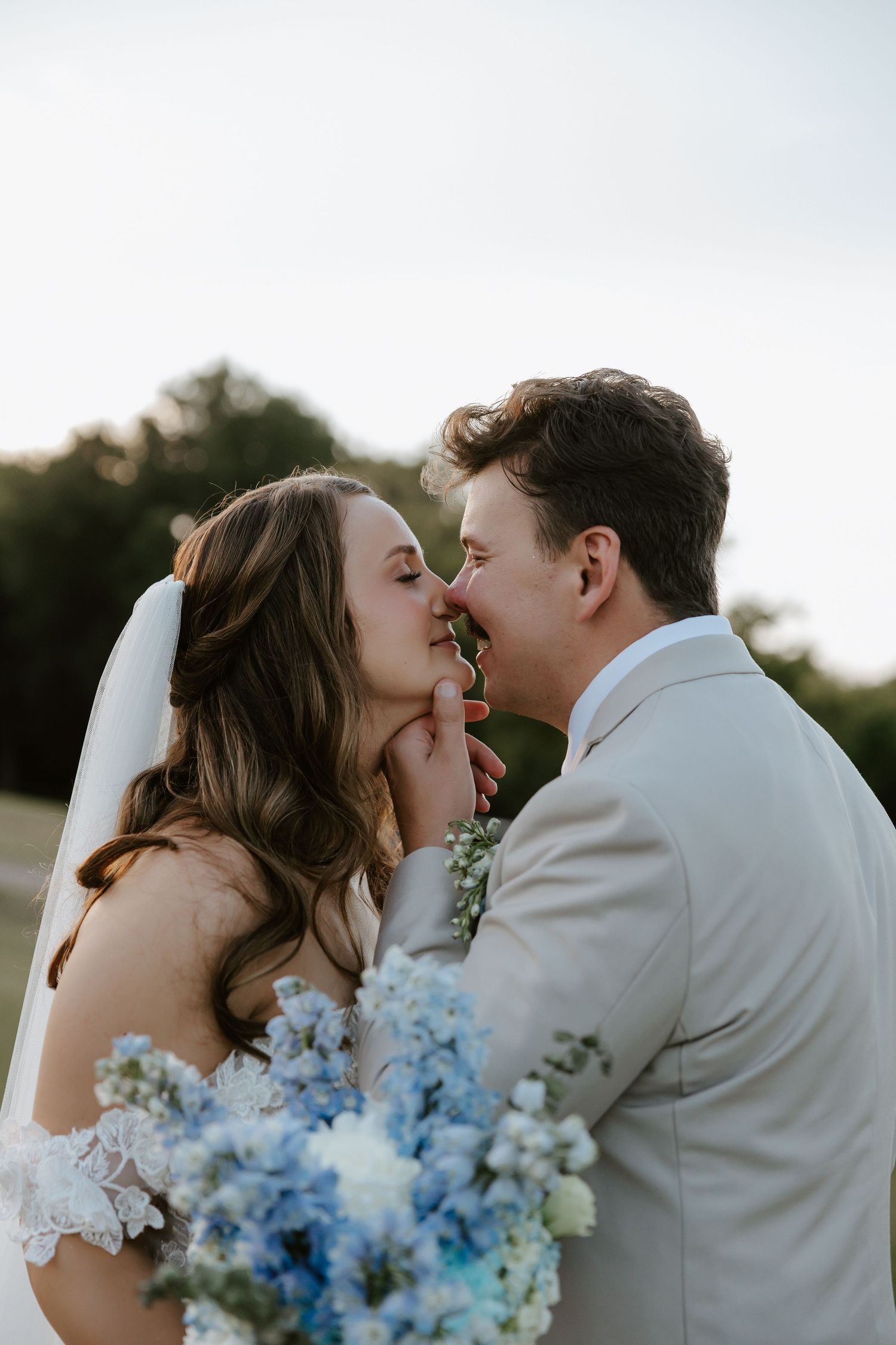 Newlyweds share intimate kisses while holding a blue and white wedding bouquet at sunset.