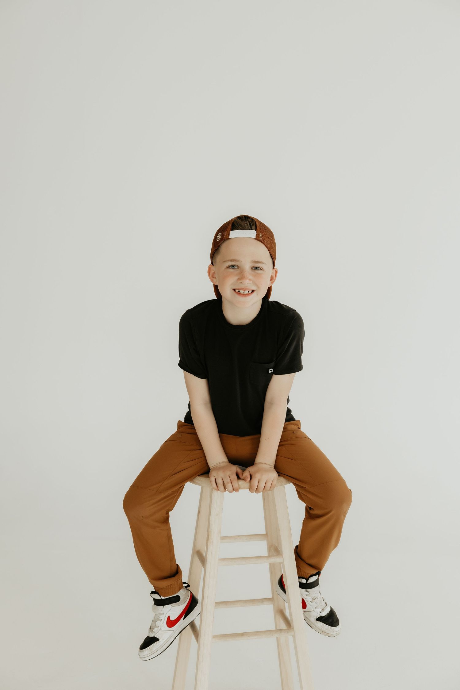 Child in casual outfit sits on wooden stool wearing sneakers against white background.