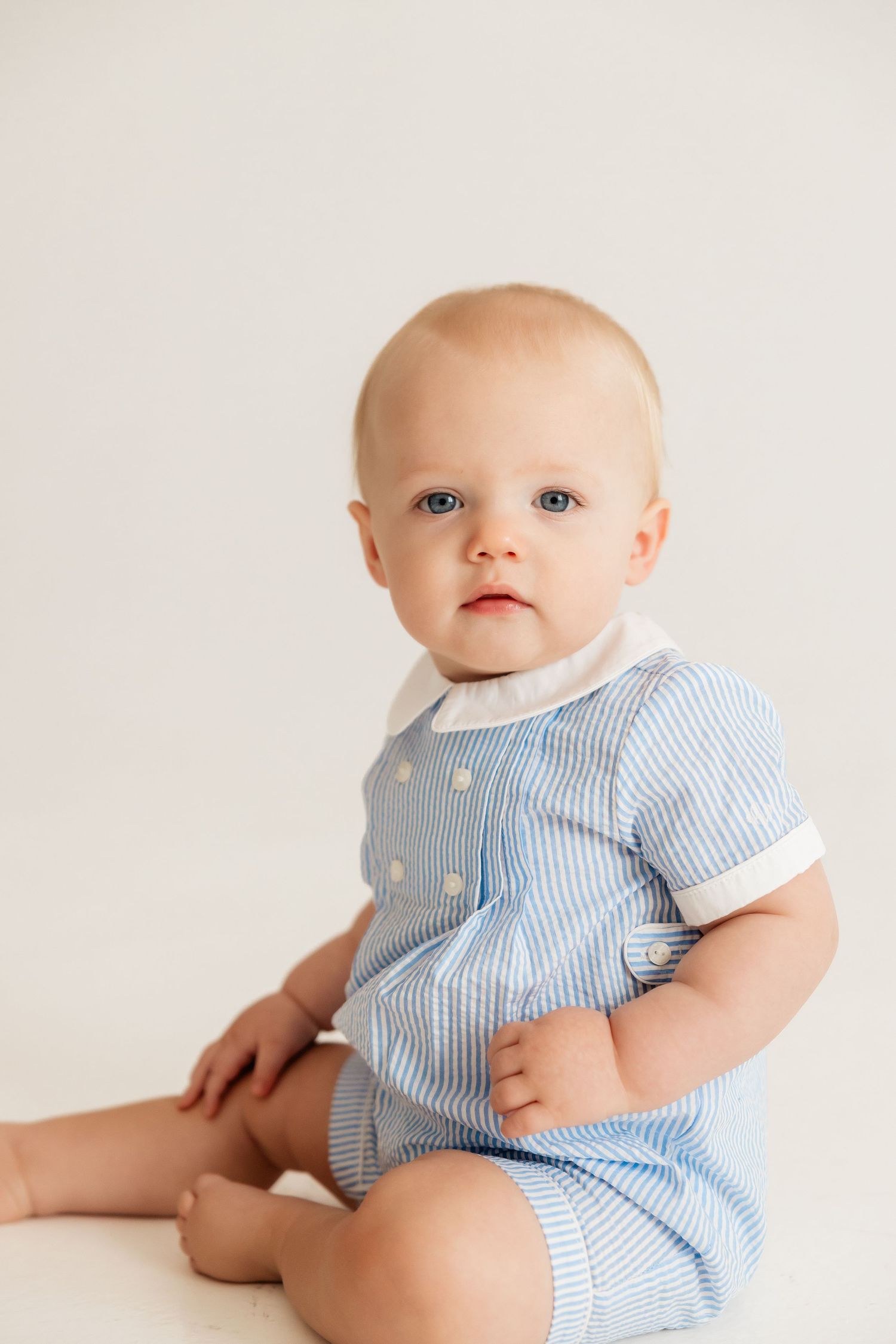 Baby wearing blue and white striped romper sits against white background.