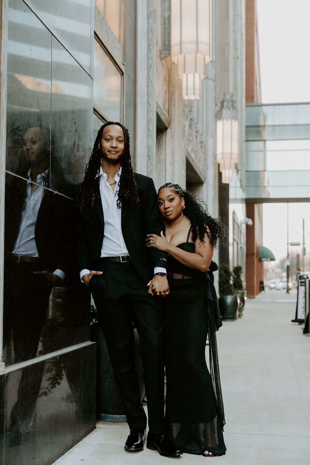 Professional couple in formal business attire posing against modern glass building exterior.