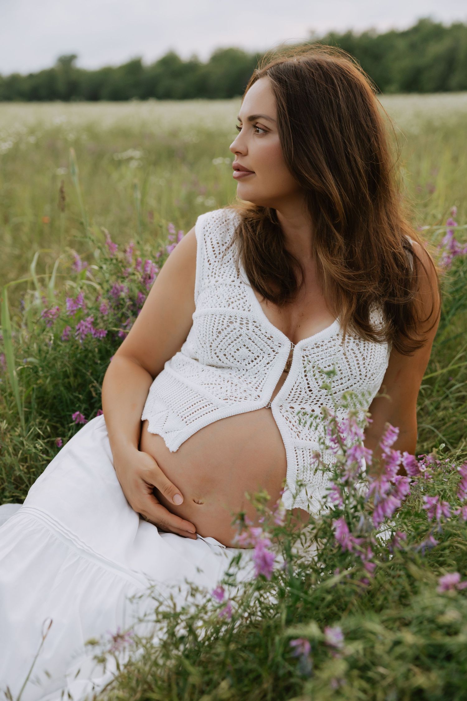 A maternity portrait in a white lace dress surrounded by purple wildflowers in a summer field.