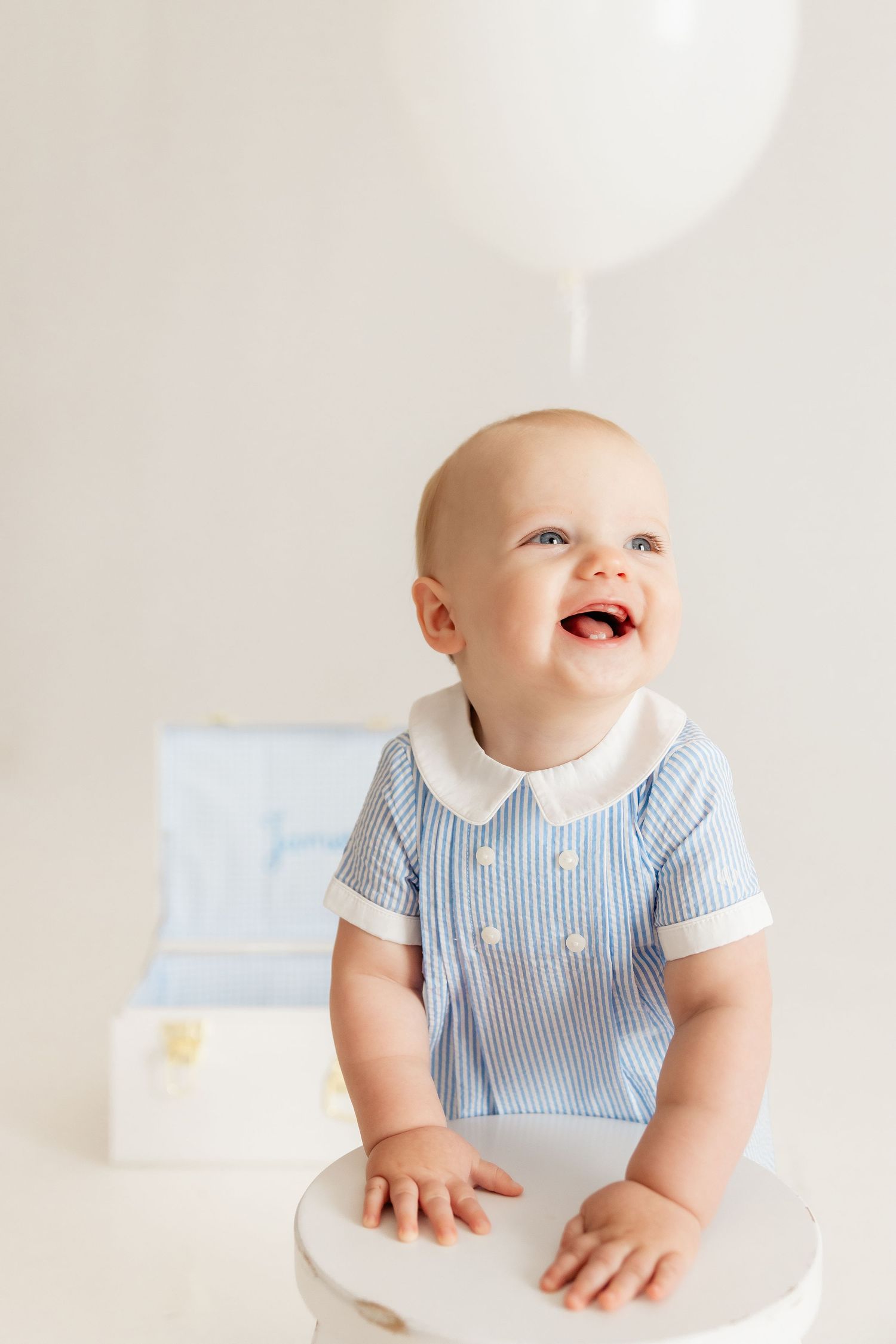 Smiling baby in blue striped outfit stands at white stool with balloon overhead.