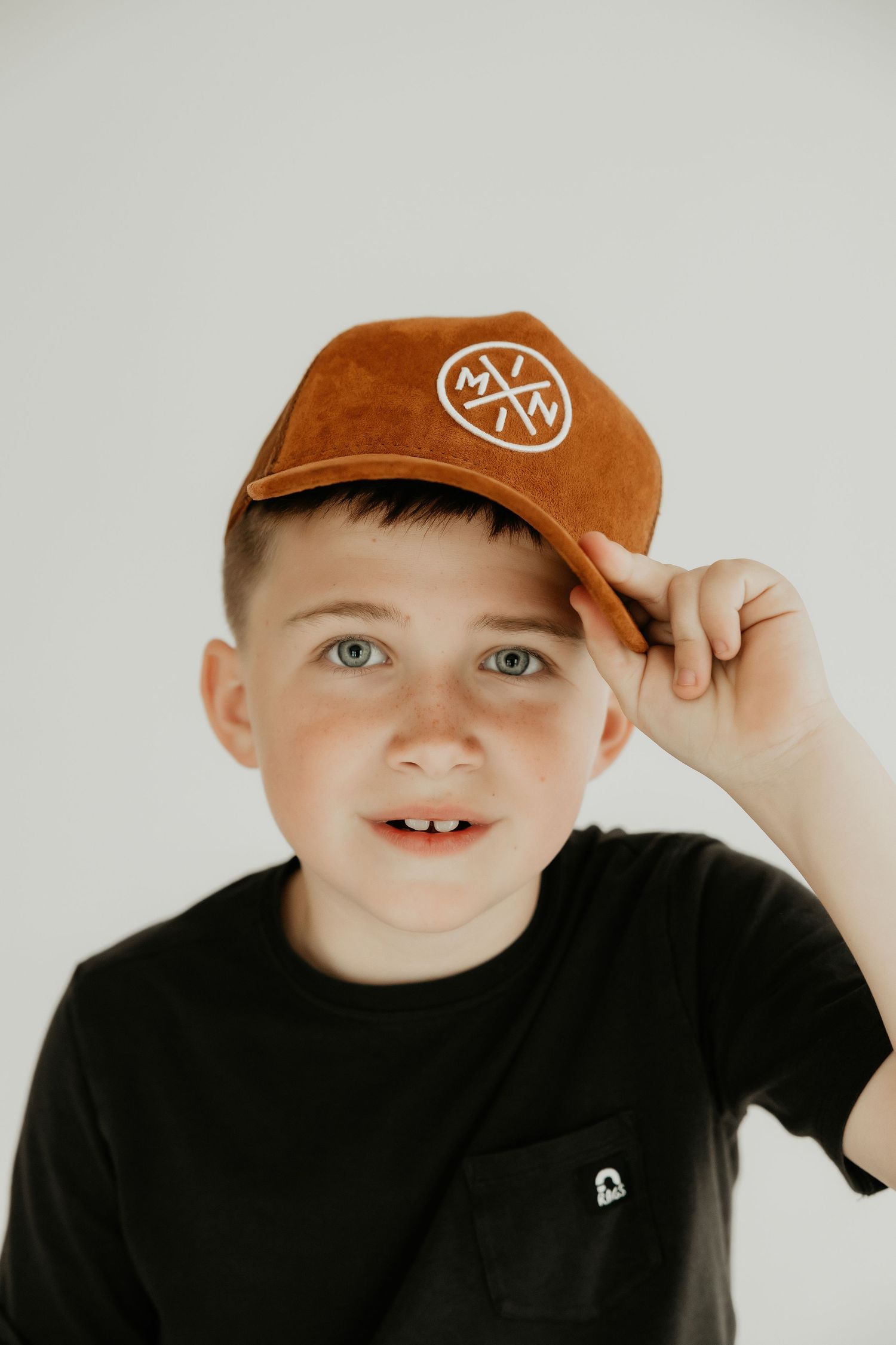 Child in black t-shirt and brown cap with circular logo smiles at camera against white backdrop.