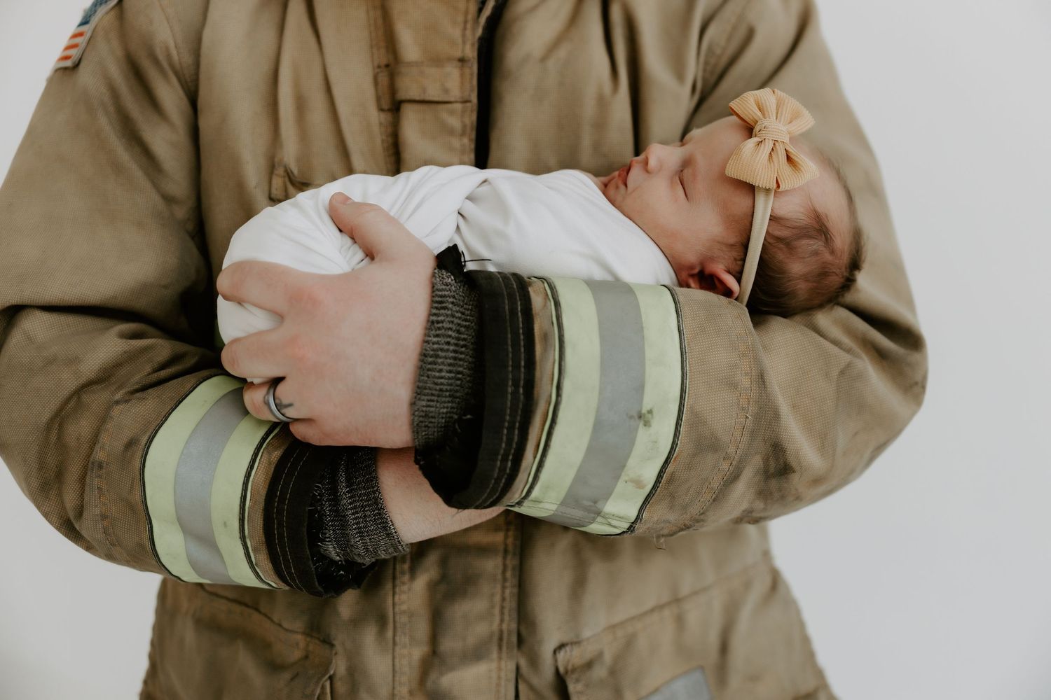 A sleeping newborn wrapped in white is cradled in firefighter turnout gear.