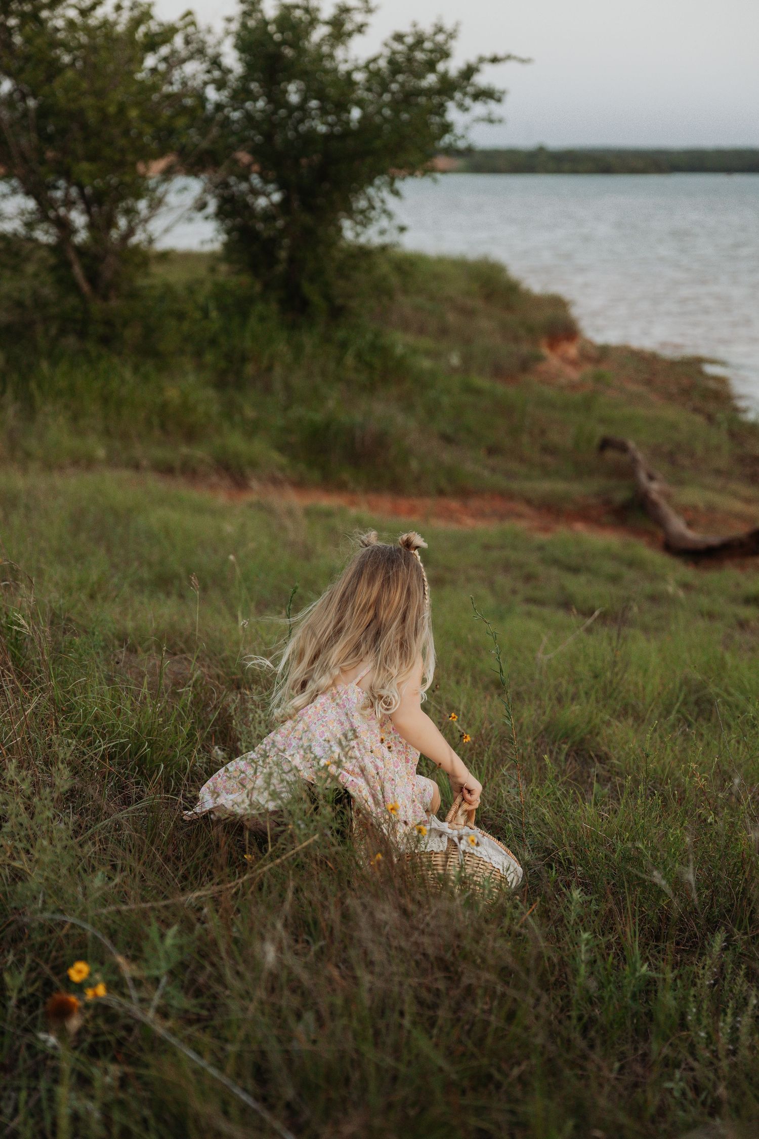 Woman in floral dress walking through wildflowers near a lake at sunset.