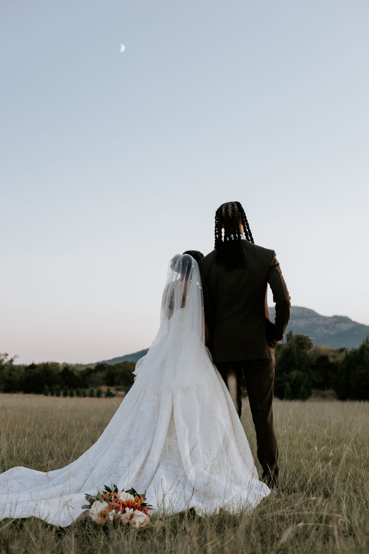 Silhouetted wedding couple in field with mountain backdrop at dusk.