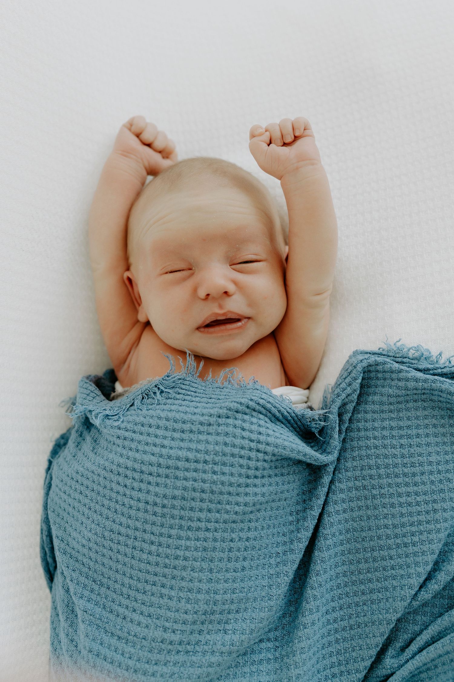 Newborn baby stretches and yawns while wrapped in a blue textured blanket.