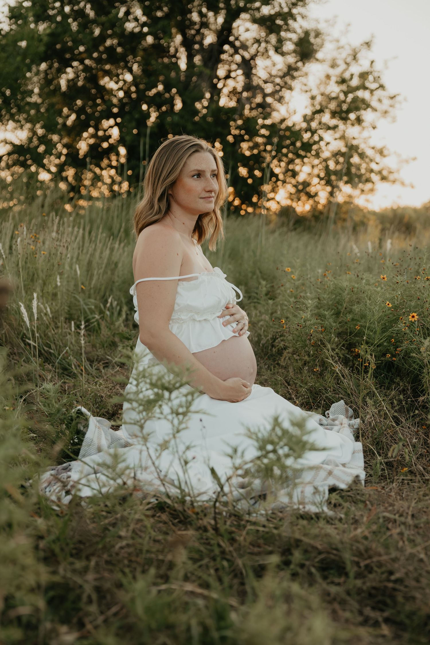 A woman in a white dress sits gracefully in a sunlit field during golden hour.