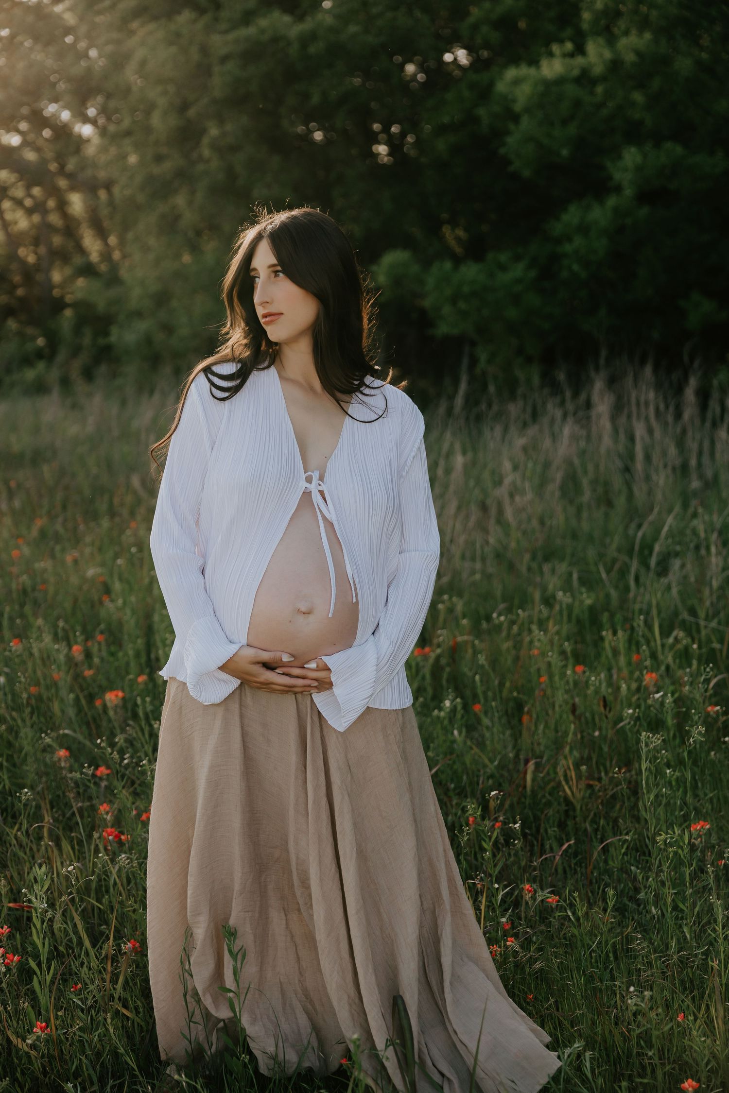 Woman in white blouse and beige dress poses gracefully in a wildflower meadow at sunset.