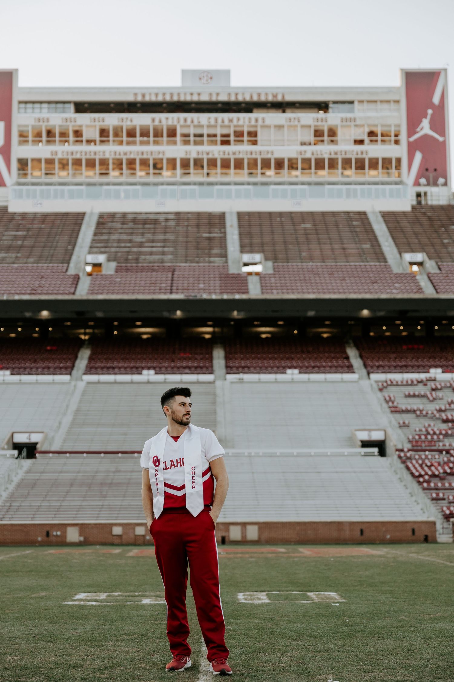 Graduate in red and white attire poses on football field at University of Oklahoma stadium.