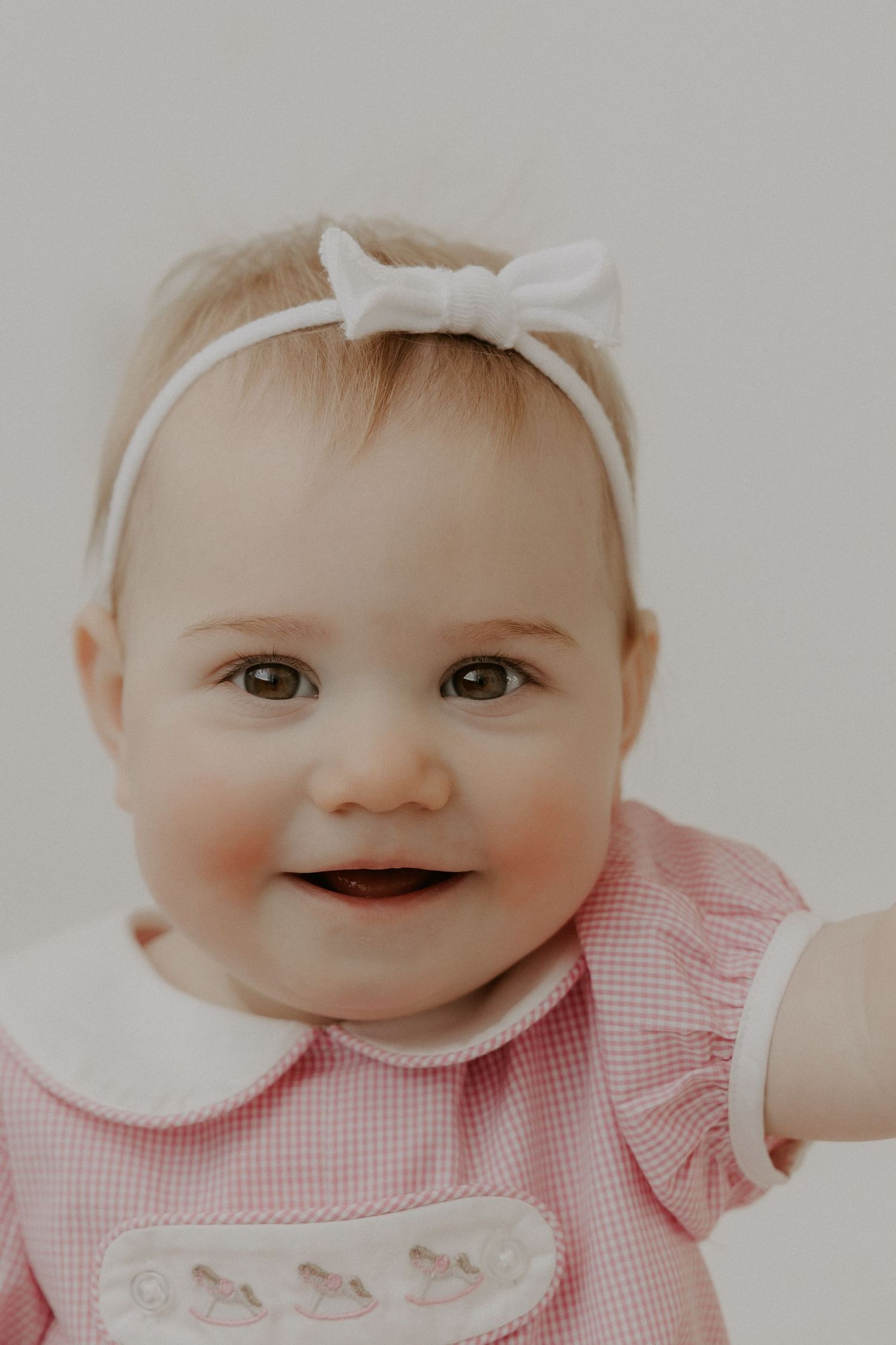 Baby in pink gingham dress and white bow headband smiling.
