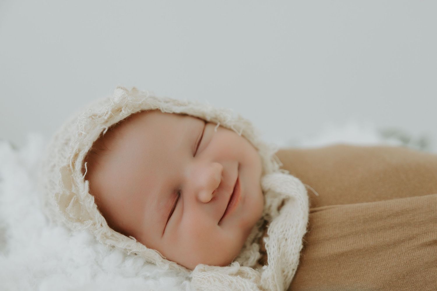 Newborn baby sleeping peacefully in cream colored wrap against light background.