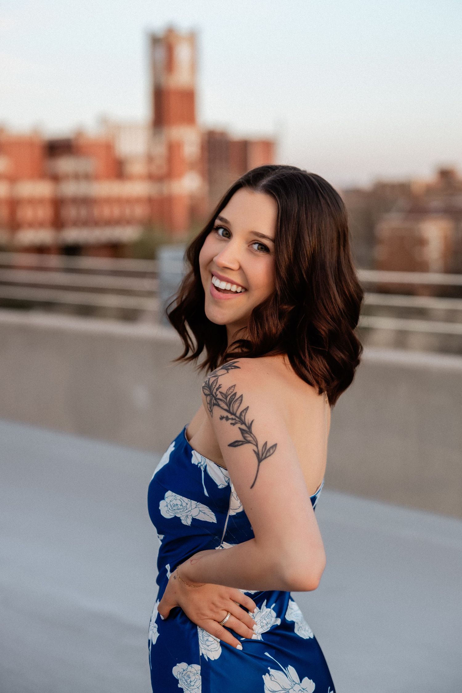 Woman in blue floral dress poses on rooftop with cityscape in background.