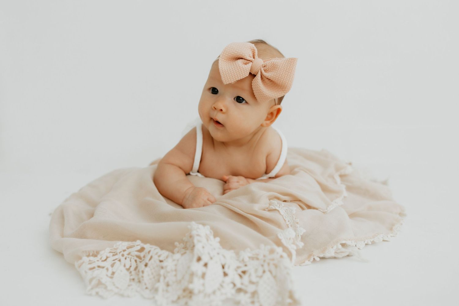 Baby in delicate lace dress lying on cream-colored fabric.