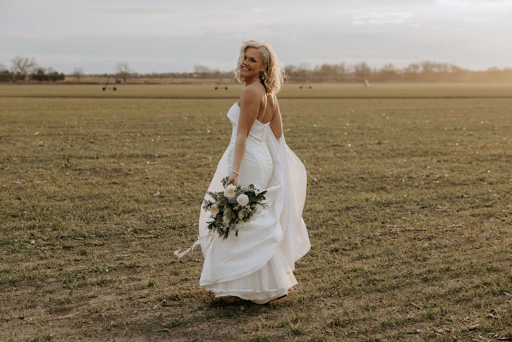 A bride in a flowing white dress walks through a golden field at sunset holding a bouquet.