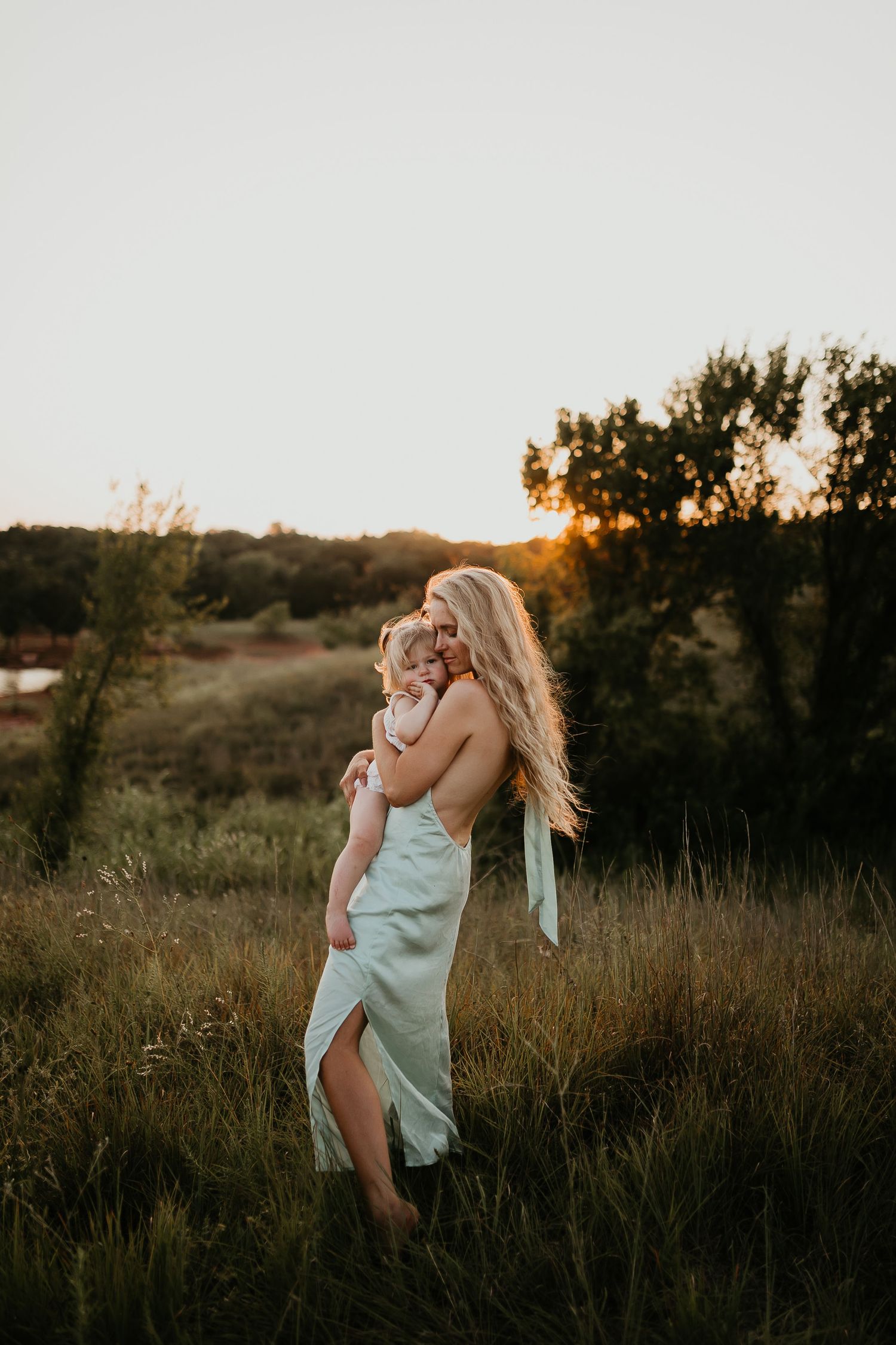 Woman in a flowy dress holding a child in a grassy field at golden hour.