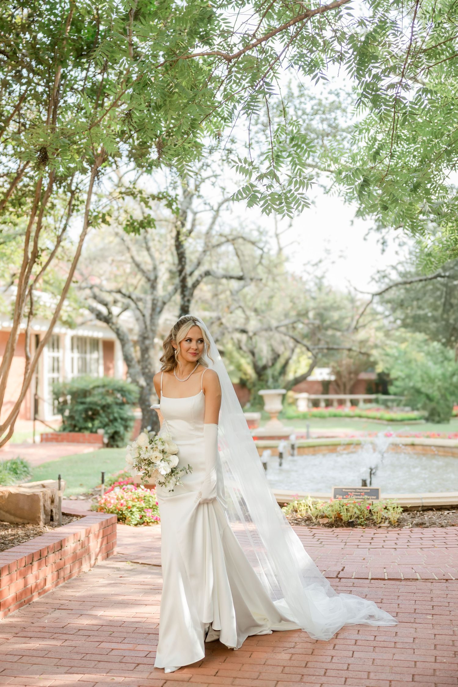 A bride in a sleek white wedding dress poses on a brick pathway surrounded by lush garden scenery.
