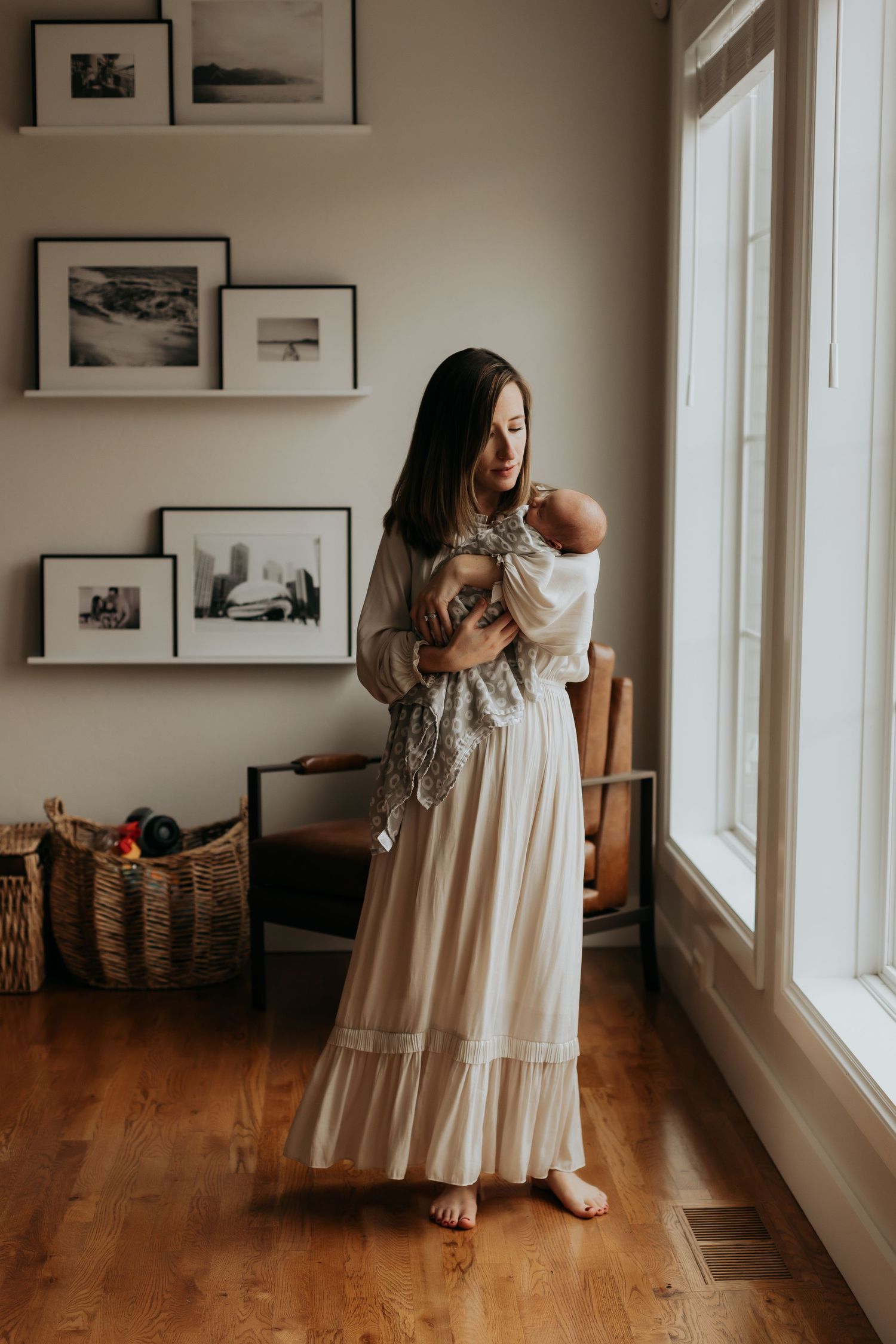 Woman in flowing white dress holds baby while standing by window with framed photos.