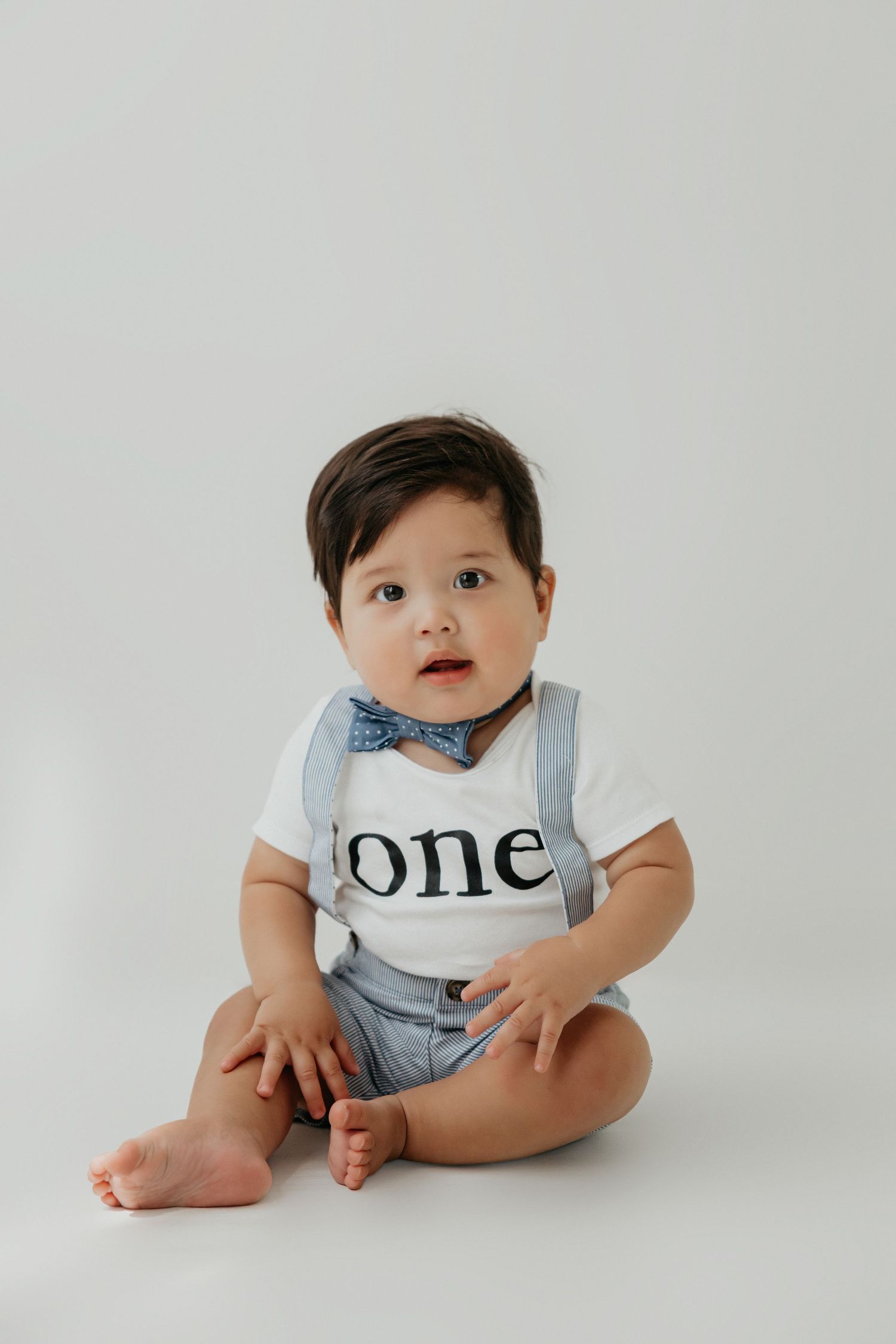 Baby wearing a 'one' shirt sits on white background showing adorable expression.