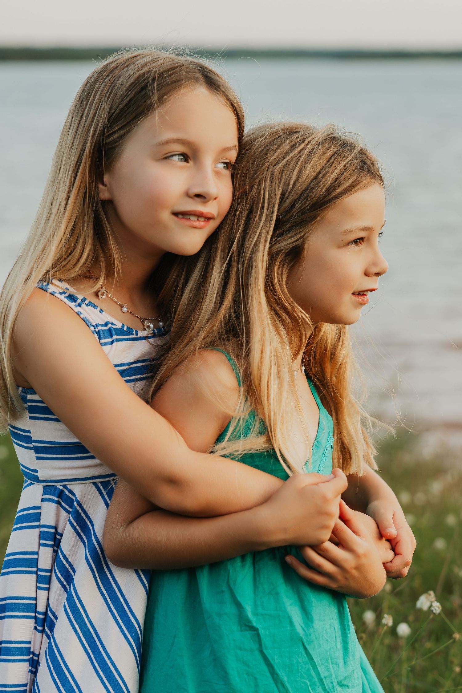 Sisters in blue and green dresses sharing a tender moment by the water.