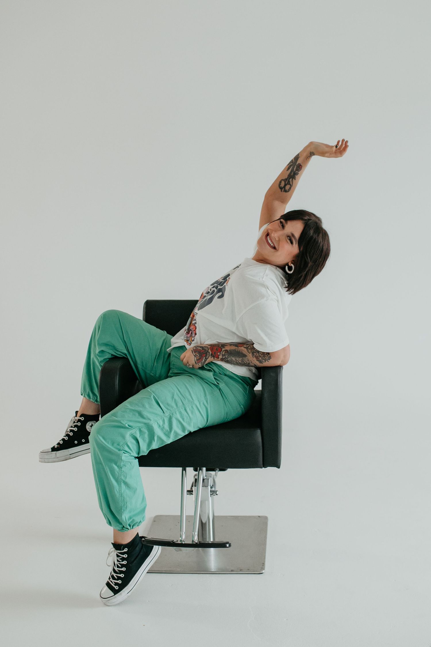 Person in green pants and graphic tee poses playfully on black chair.