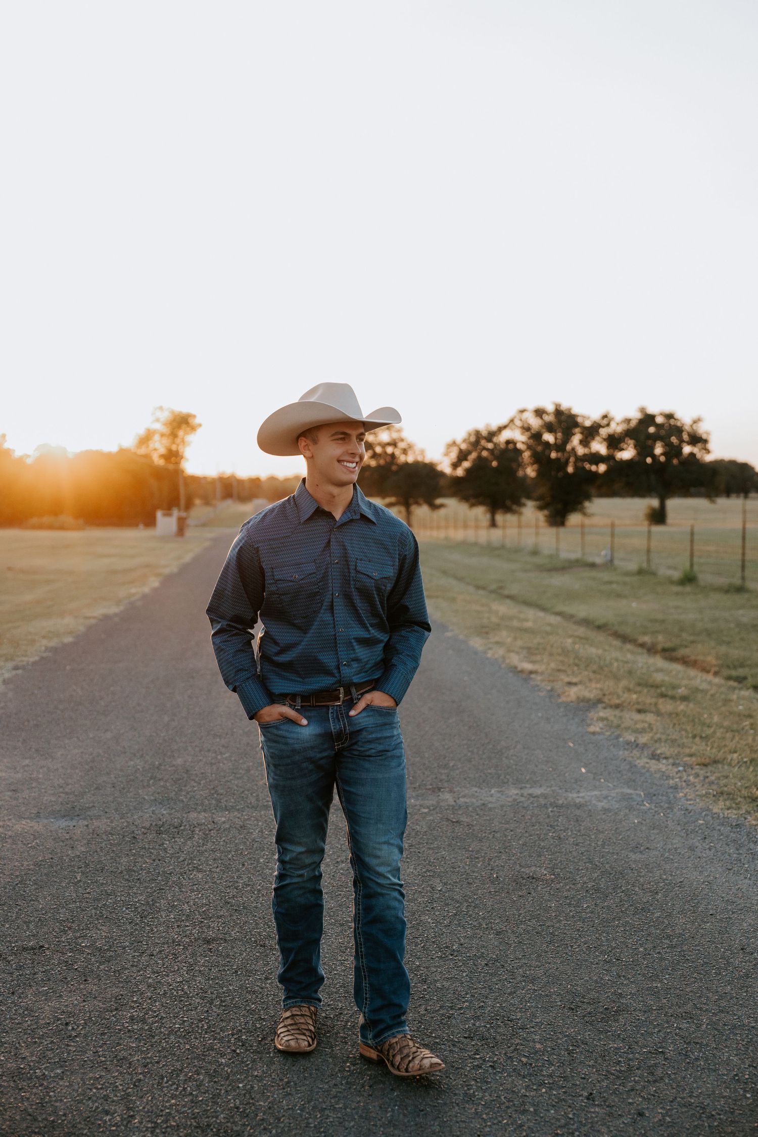 Person in western attire poses on rural road at sunset.