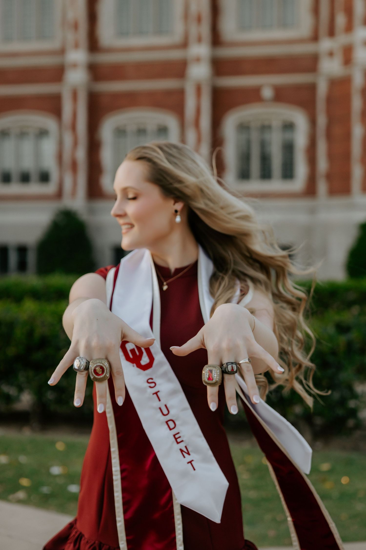 Graduate in crimson attire twirling with stole and sash outdoors.